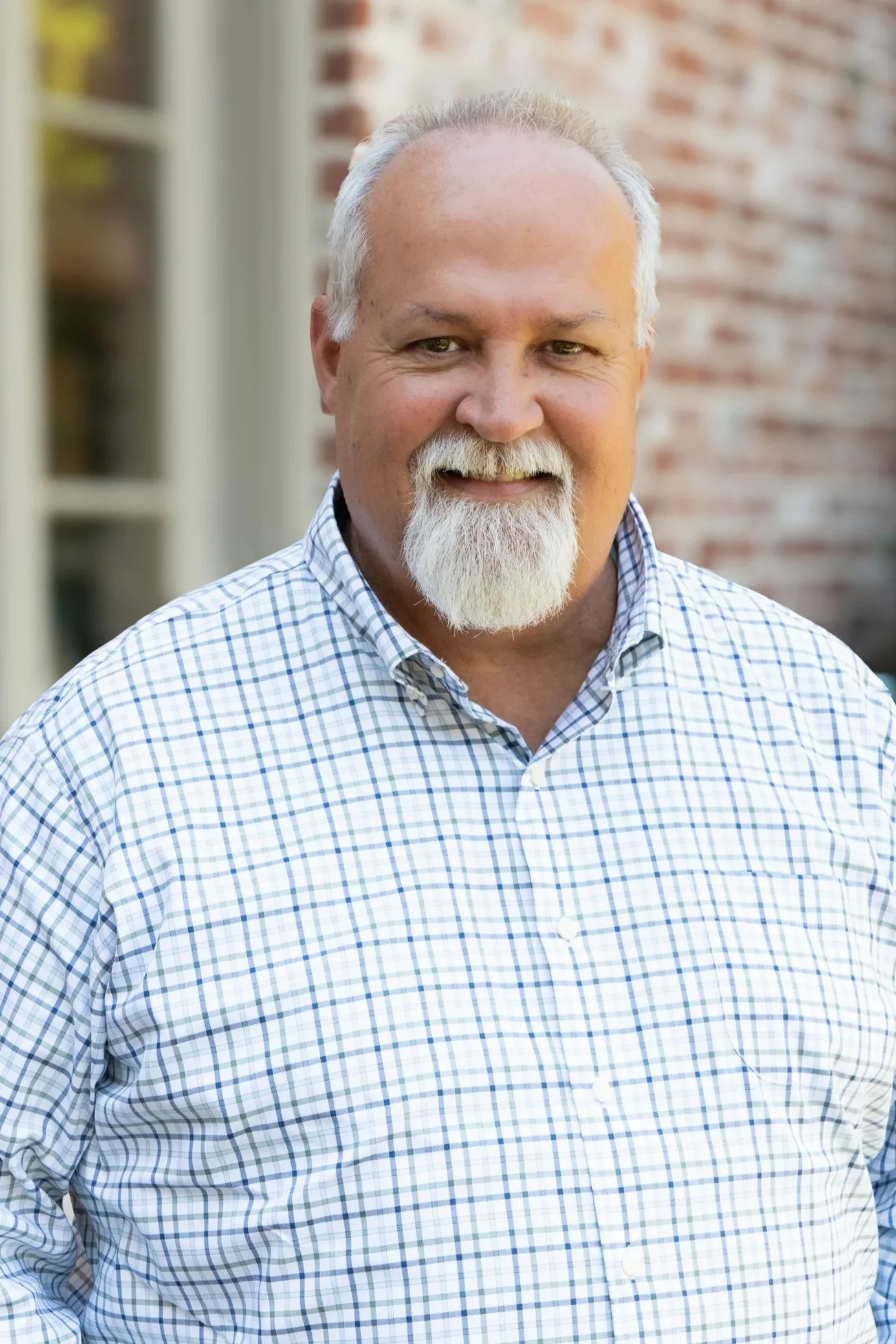 Portrait of an older man with gray hair and beard, wearing a checkered shirt, standing outdoors with a blurred brick wall background.