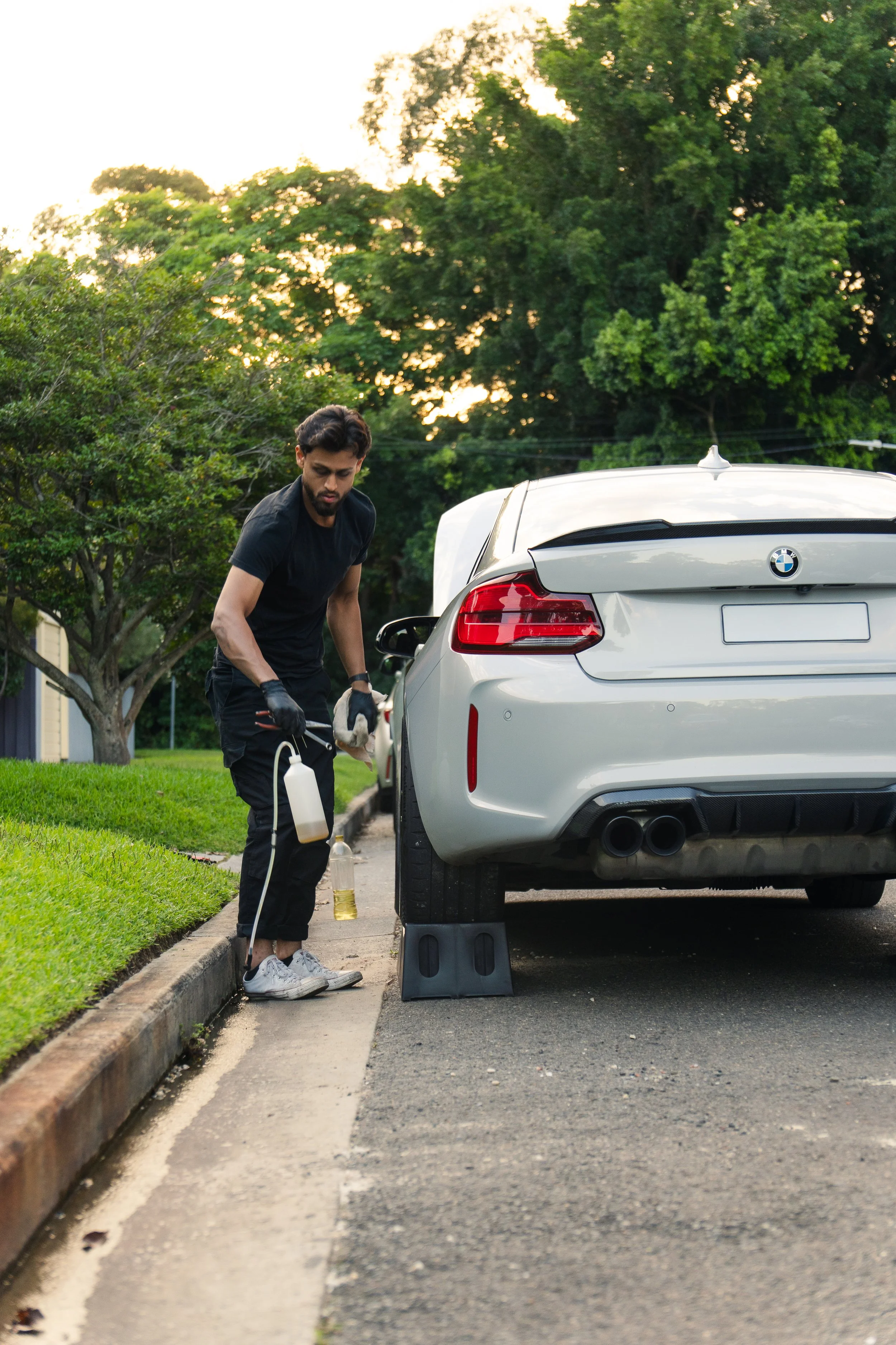 A man in black clothing wearing gloves working underneath a silver BMW parked on the street during sunset, with trees and houses in the background.