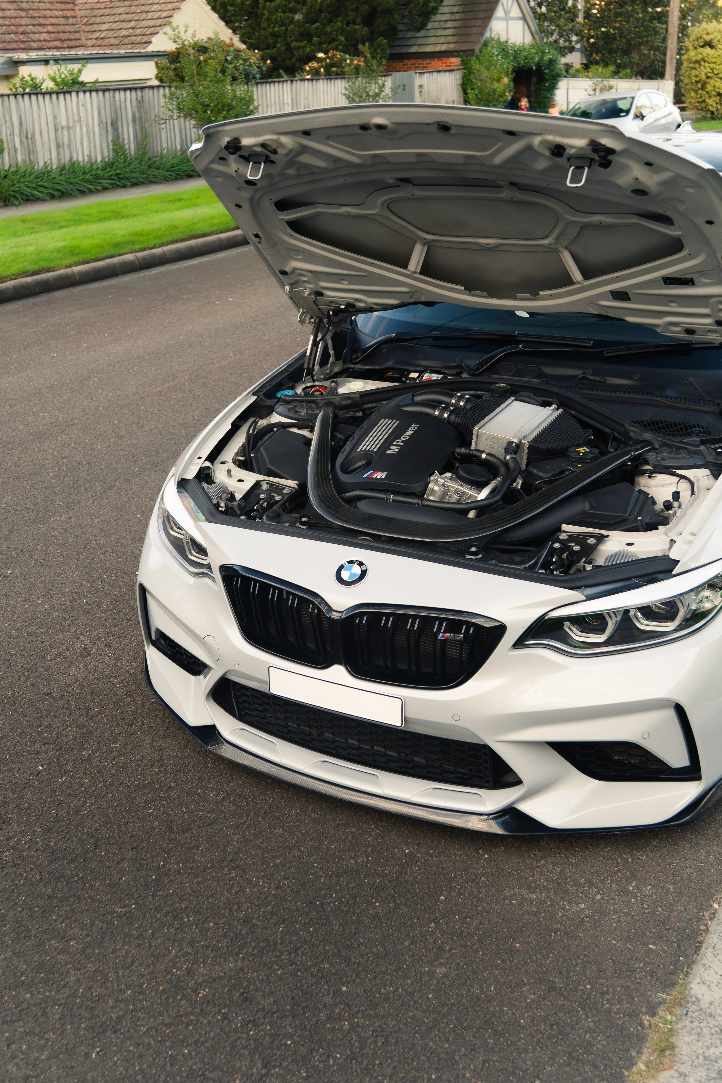 A white BMW car with its hood open, exposing the engine bay, parked on a residential street with houses and greenery in the background.