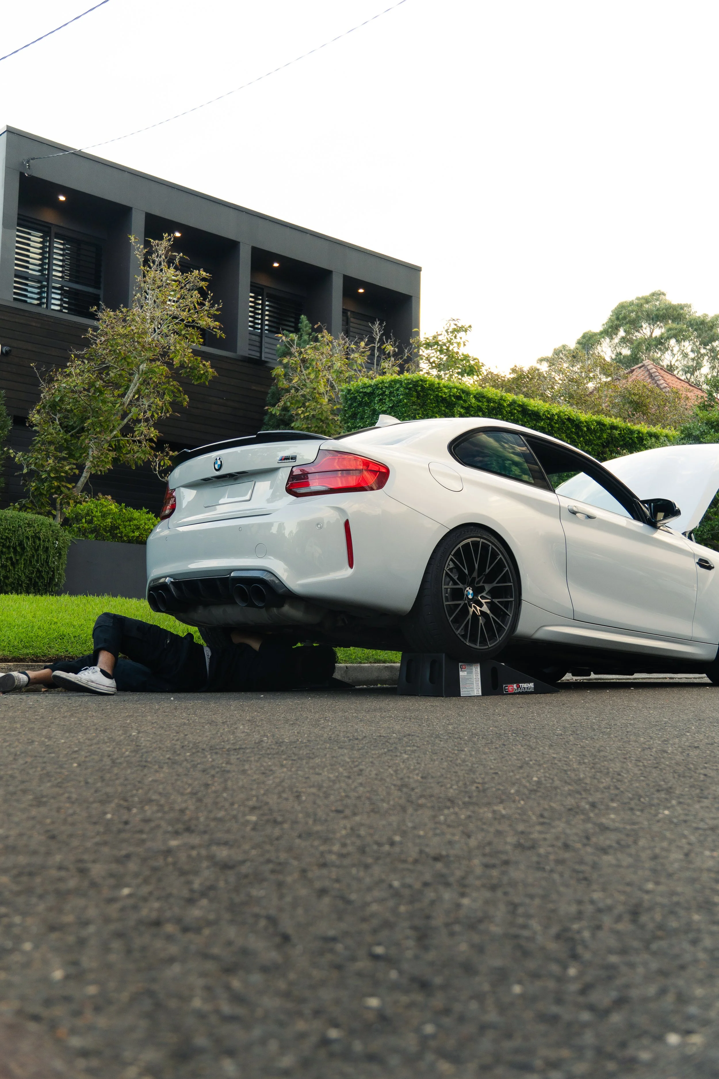 A person working underneath a white BMW M2 car that is elevated on stilts on a driveway, with a modern house and trees in the background.