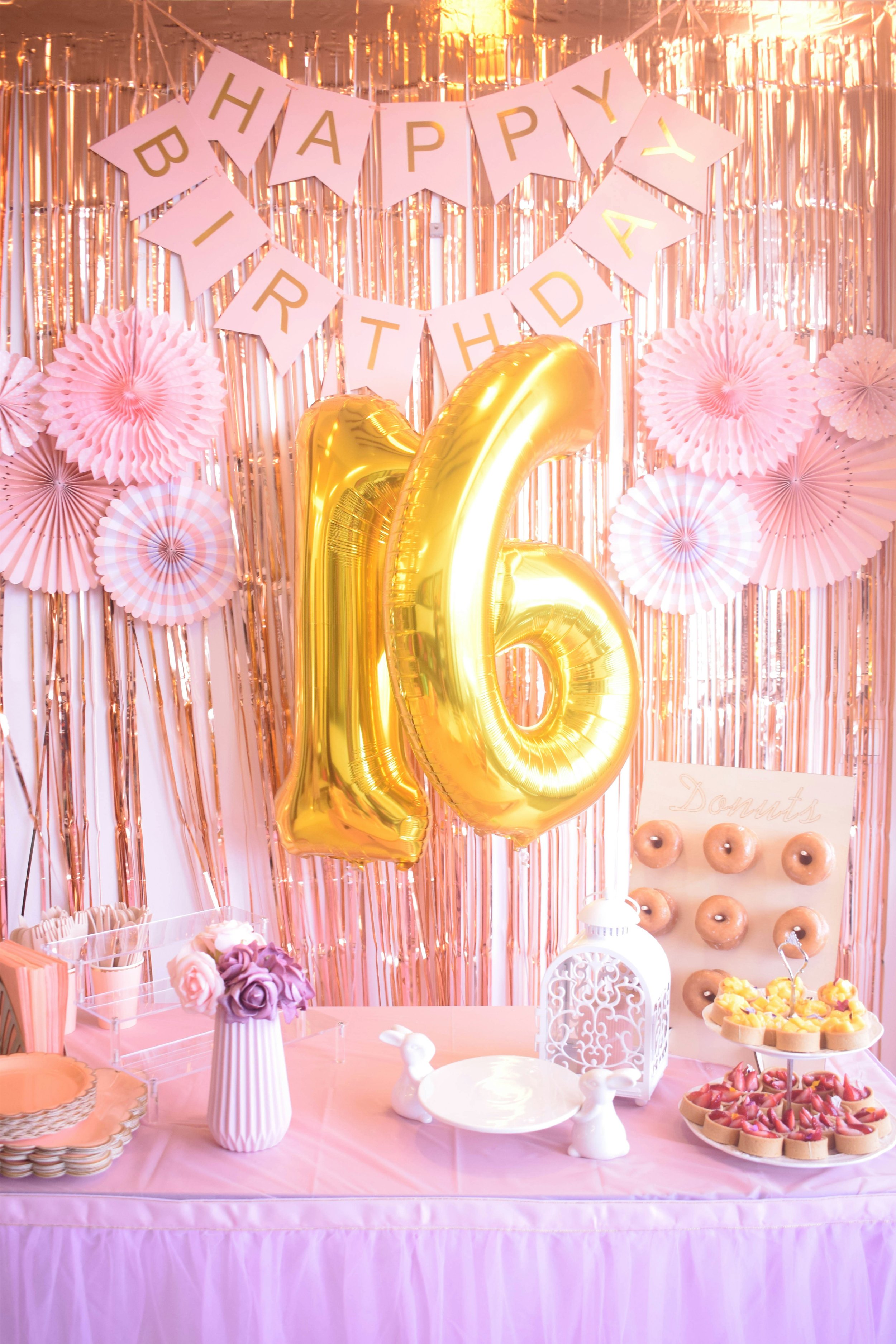 Decorative birthday celebration setup with pink and gold theme. Pink paper fans and metallic balloons spell out 'Happy Birthday' and display the number '16'. The table includes various desserts like donuts, cupcakes, and fruit tarts, along with pink roses in a white vase. A pink tablecloth covers the table, and background features pink and gold streamers.
