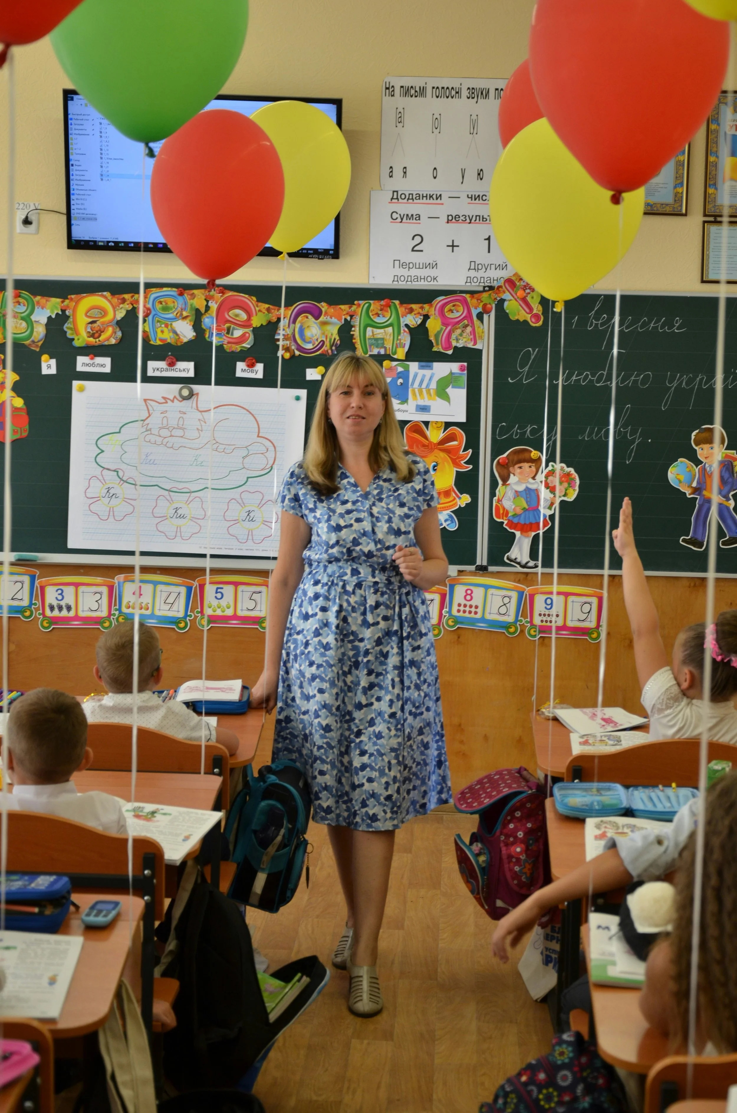 A classroom filled with students sitting at desks, with a woman standing at the front near a blackboard. Balloons are floating in the classroom, and colorful decorations hang on the walls. The children are engaged, some raising hands, and the classroom is decorated for a celebration.