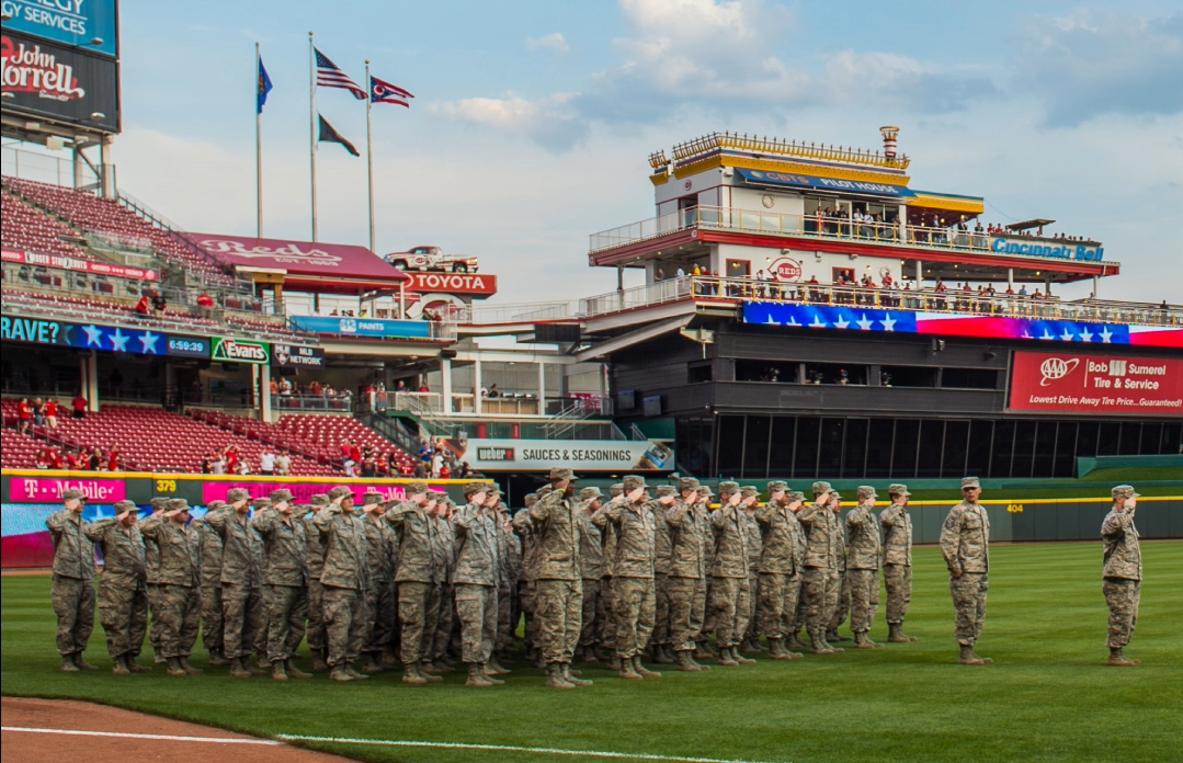 Group of soldiers in camouflage uniforms saluting on the baseball field at a stadium with flags flying above and spectators in the stands.