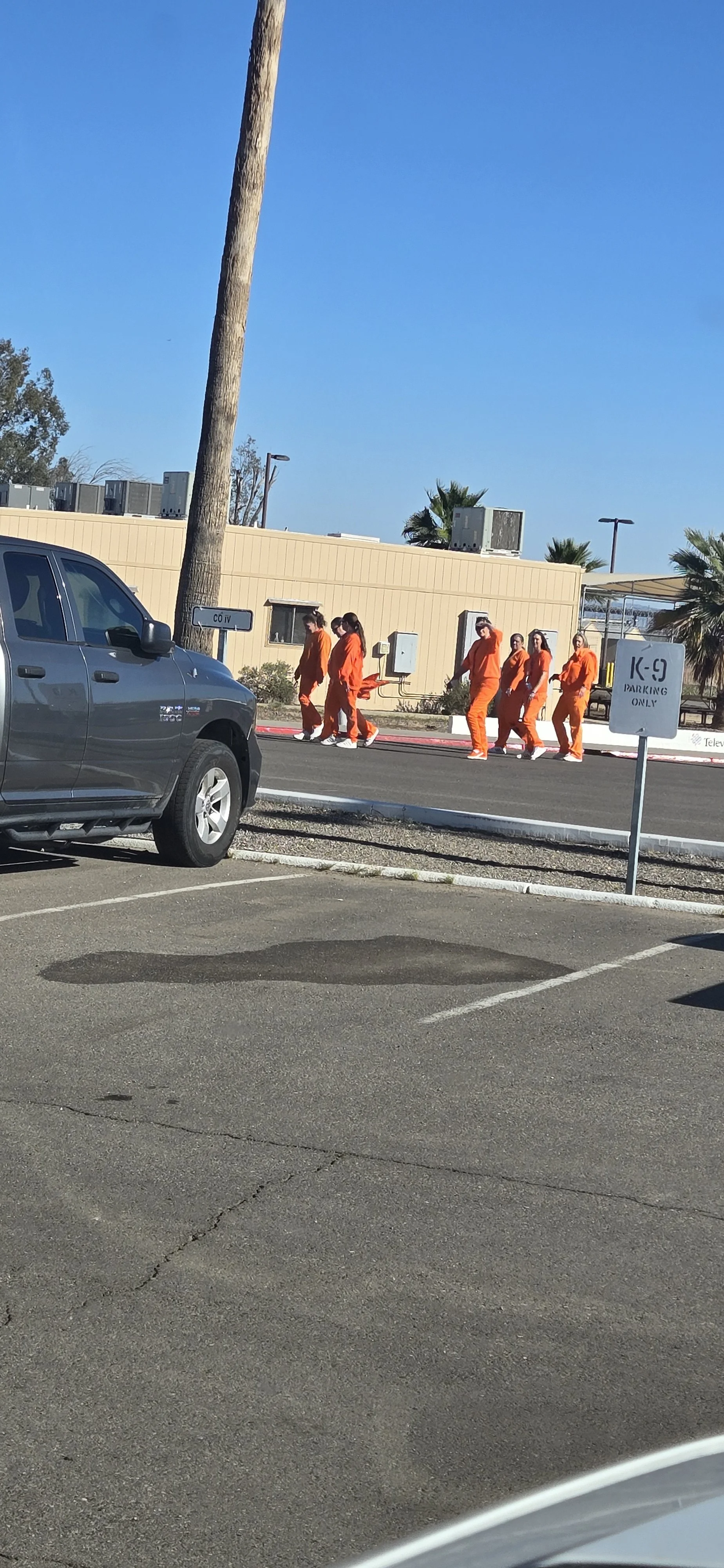 Group of people dressed in orange uniforms walking in a parking lot near a beige building with palm trees and a clear blue sky.