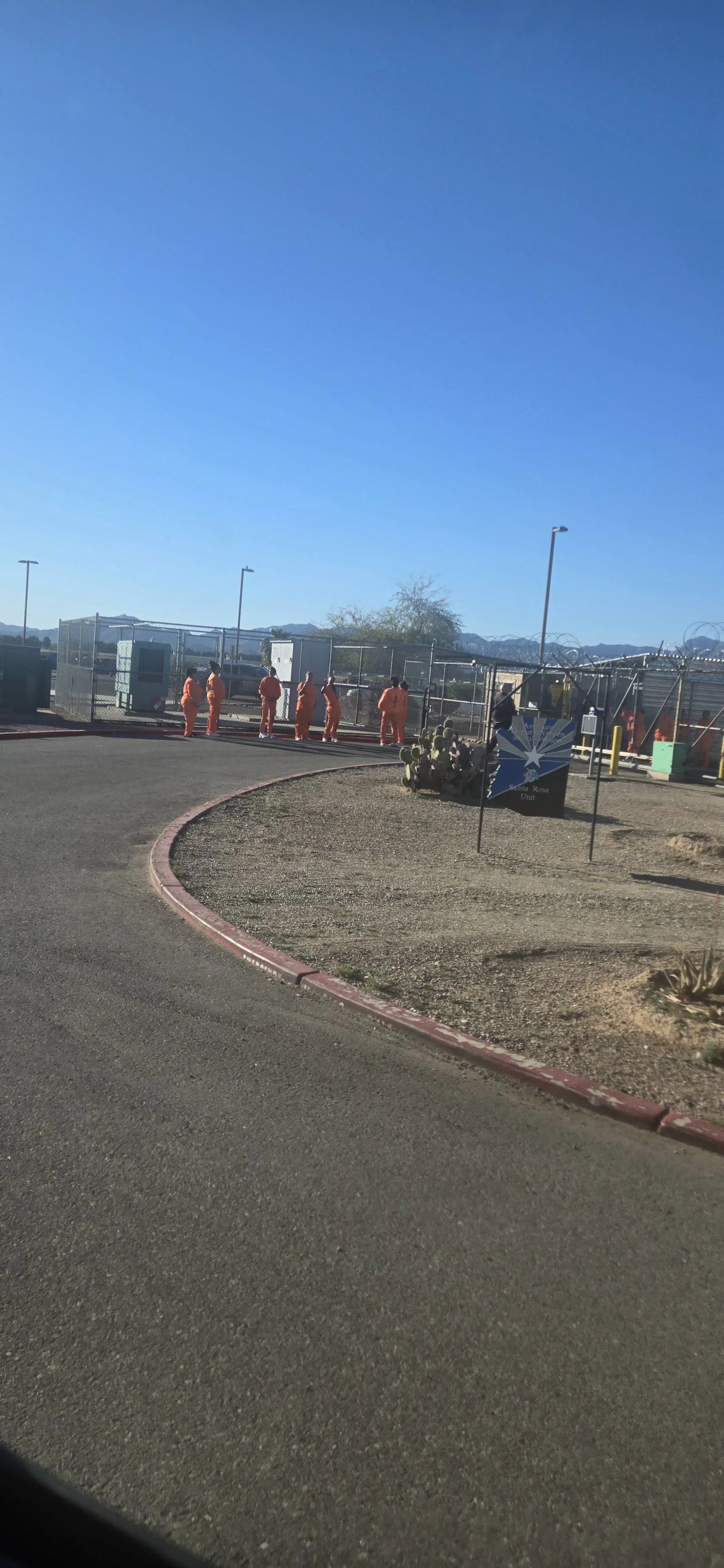 Several inmates in orange jumpsuits are standing near a fenced area at a prison or detention center, with mountains in the background and desert plants nearby.