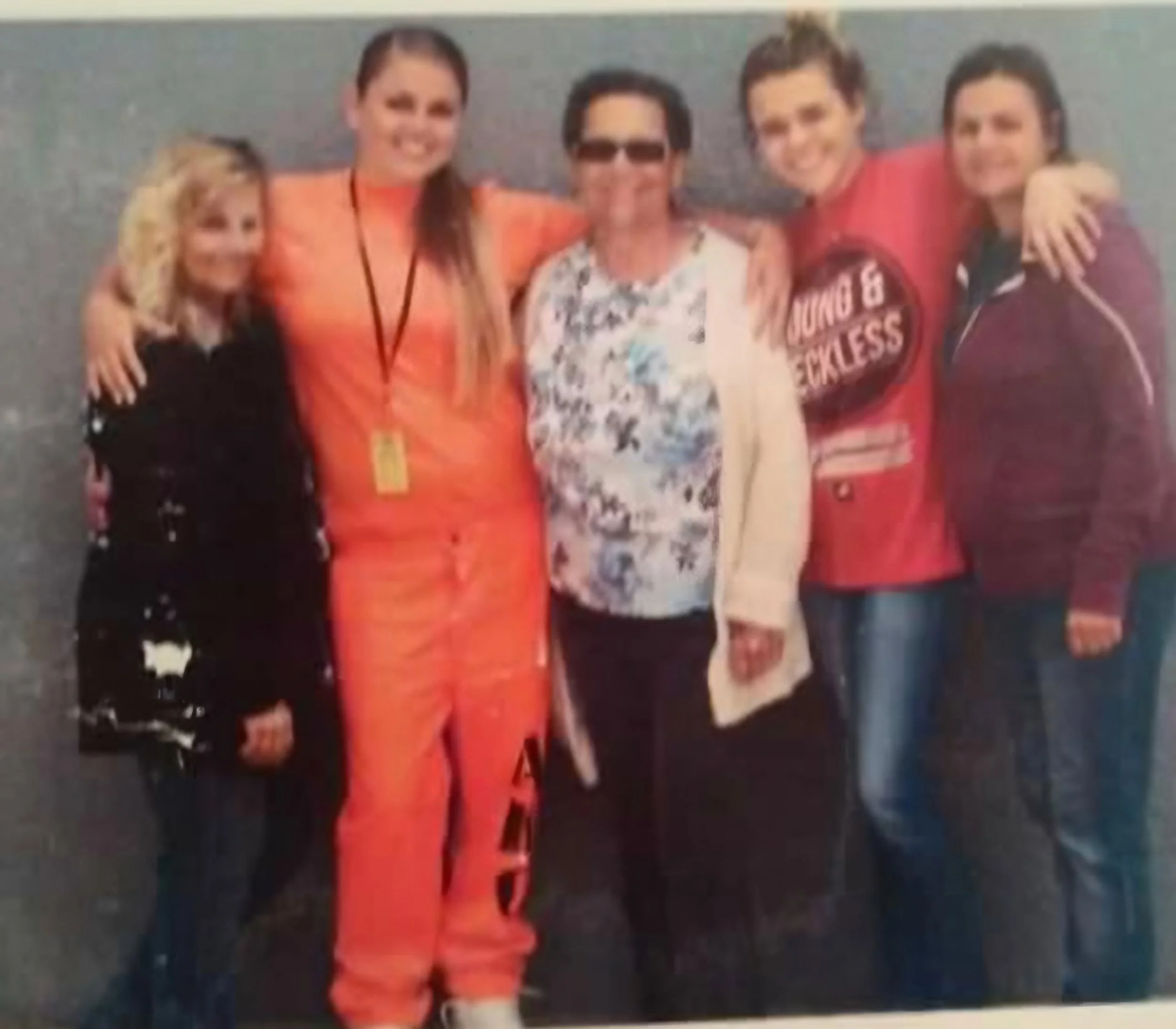 Group of five people posing together indoors, smiling. Four are women and one is a man wearing sunglasses, with the women dressed in casual clothing.