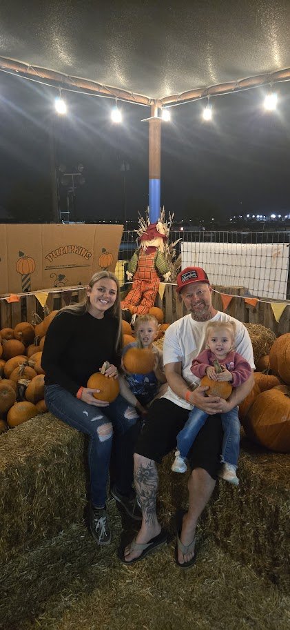 A family sits on hay bales among pumpkins at a pumpkin patch, with a Halloween scarecrow, pumpkins, and festive string lights in the background.