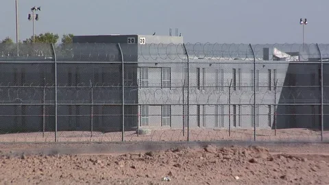 A fenced area with barbed wire, a building in the background, and a chain-link fence in the foreground.