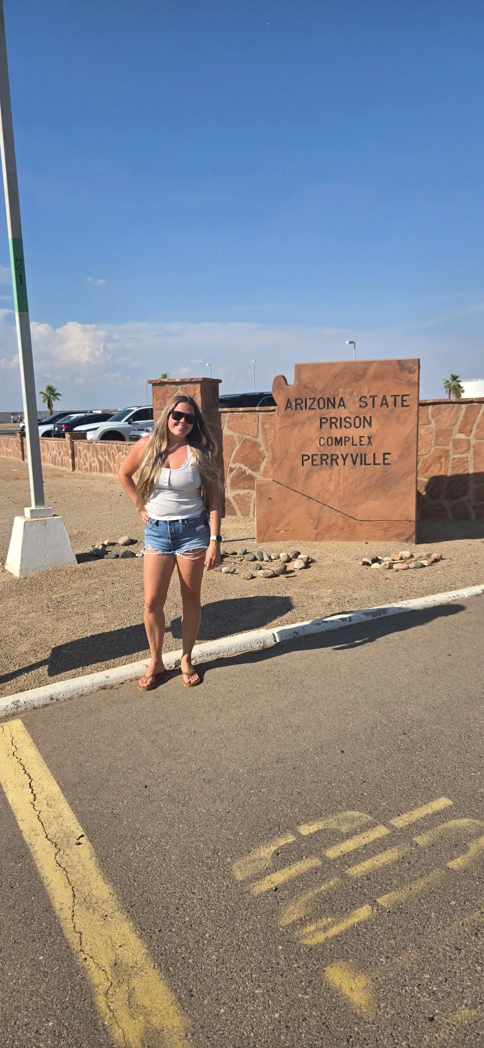 A woman wearing sunglasses, a white tank top, and denim shorts standing in front of a sign that reads 'Arizona State Prison Complex Perryville' at the prison entrance. There are parked cars and palm trees in the background under a clear blue sky.