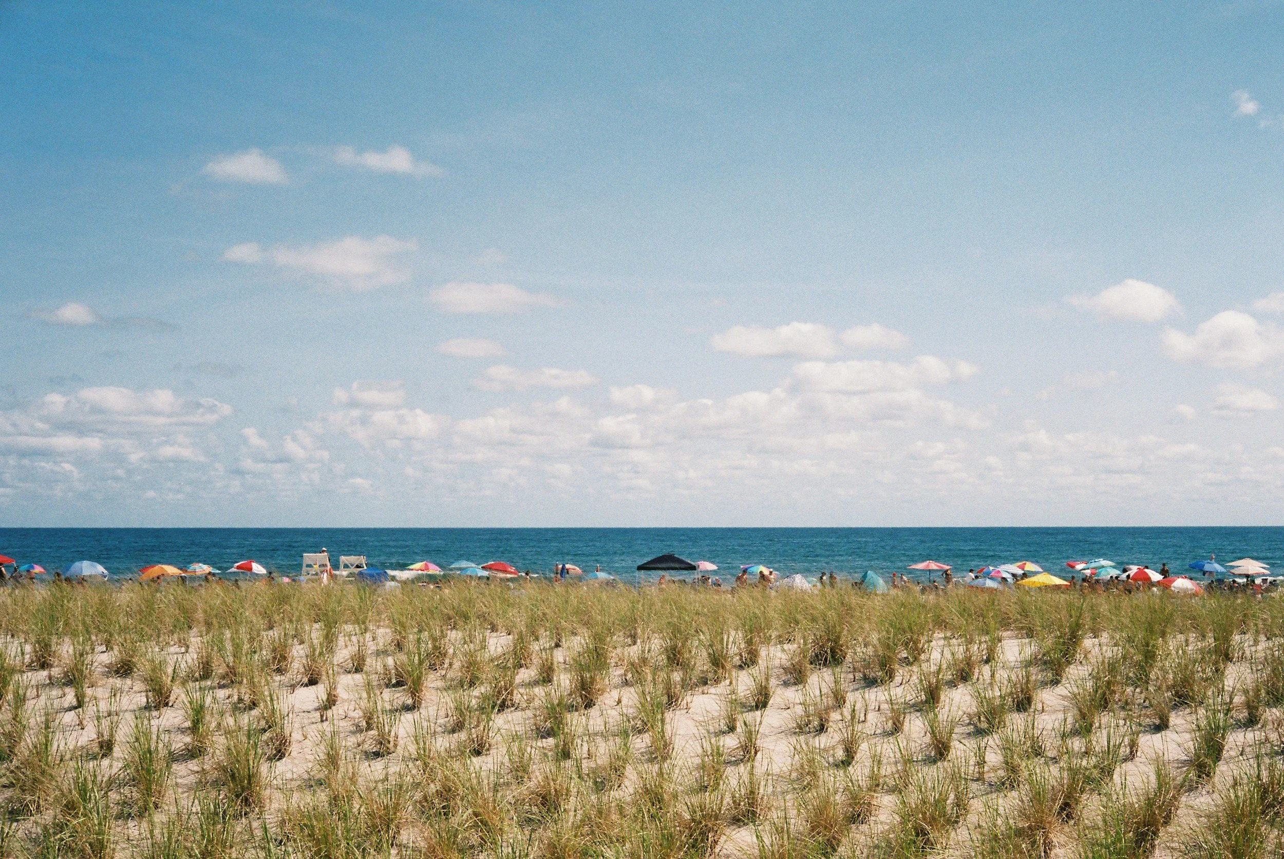 Beach scene with colorful umbrellas, grassy dunes, and the ocean in the background under a blue sky with scattered clouds.