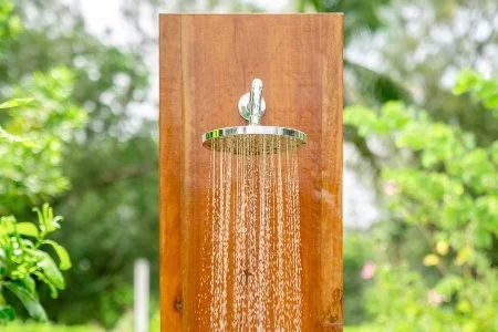 Outdoor wooden shower with water flowing from a silver showerhead, surrounded by green foliage.