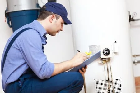 A technician in blue uniform and cap inspecting or taking notes on a clipboard next to industrial equipment or boiler.