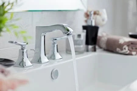 Close-up of a bathroom sink faucet with running water, with a mirror and plant in the background.
