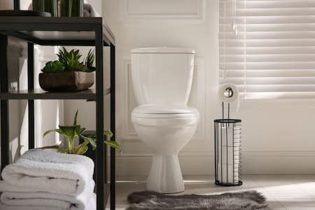 A bathroom with a white toilet, black shelving unit with potted plants and folded towels, a window with blinds, and a basket.