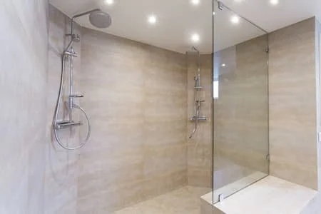 A modern, minimalistic shower area with beige tiles, two showerheads, and a glass partition.