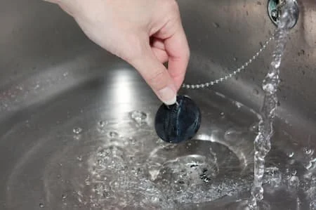 Hand holding a black sink stopper under running water in a kitchen sink.