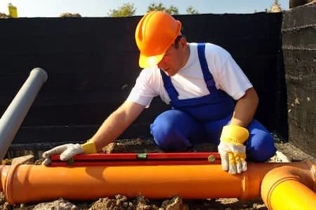 Worker wearing a yellow safety helmet and gloves, crouching and working on orange drainage pipes outdoors.