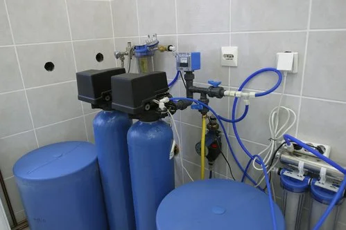 Water filtration or purification system with blue tanks, filters, and connecting tubes mounted on a wall in a utility room or laboratory setting.
