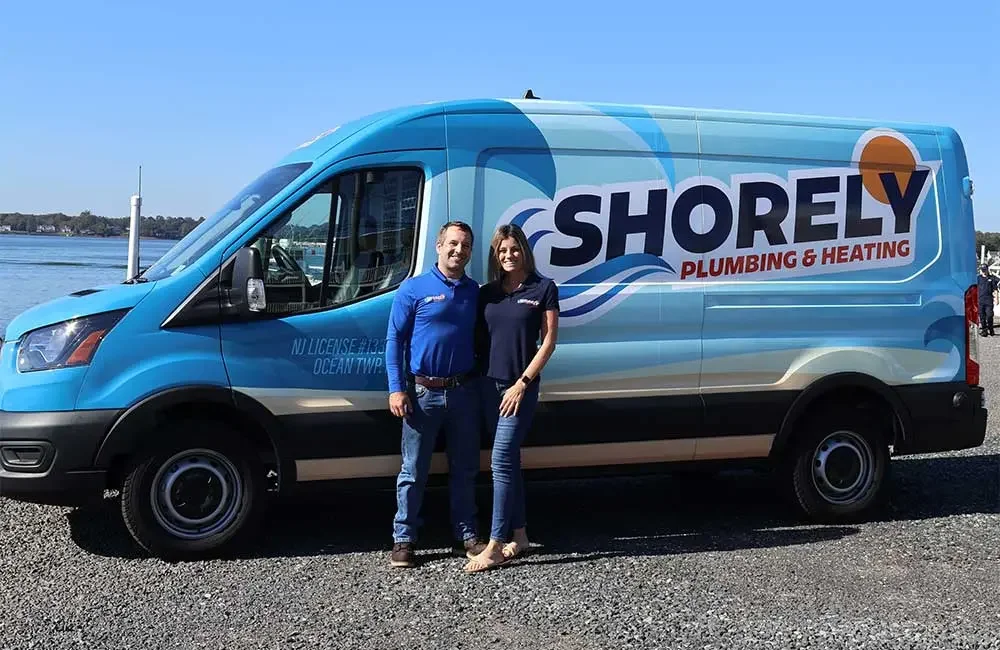 A man and woman standing in front of a Shorely Plumbing & Heating company van by the water on a clear day.
