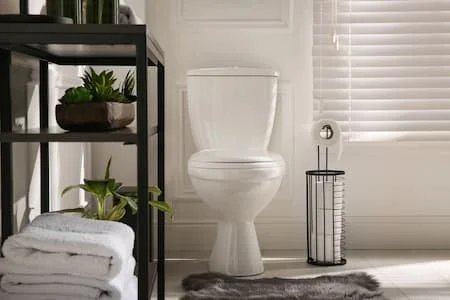 A bathroom with a white toilet, black shelving unit with plants and towels, and a brushed metal waste bin next to the toilet.