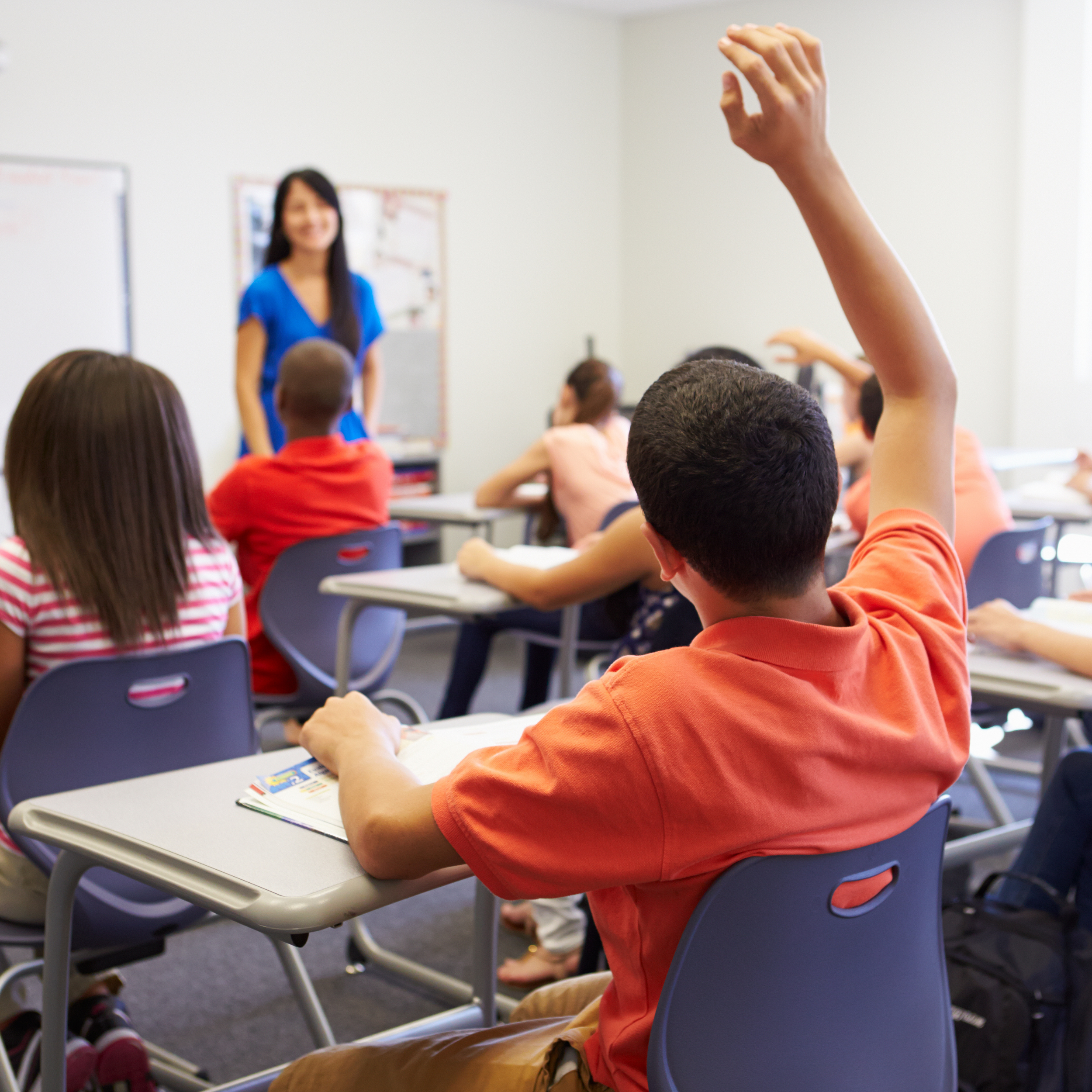 A classroom scene with students sitting at desks and a teacher standing at the front. One student has their hand raised.