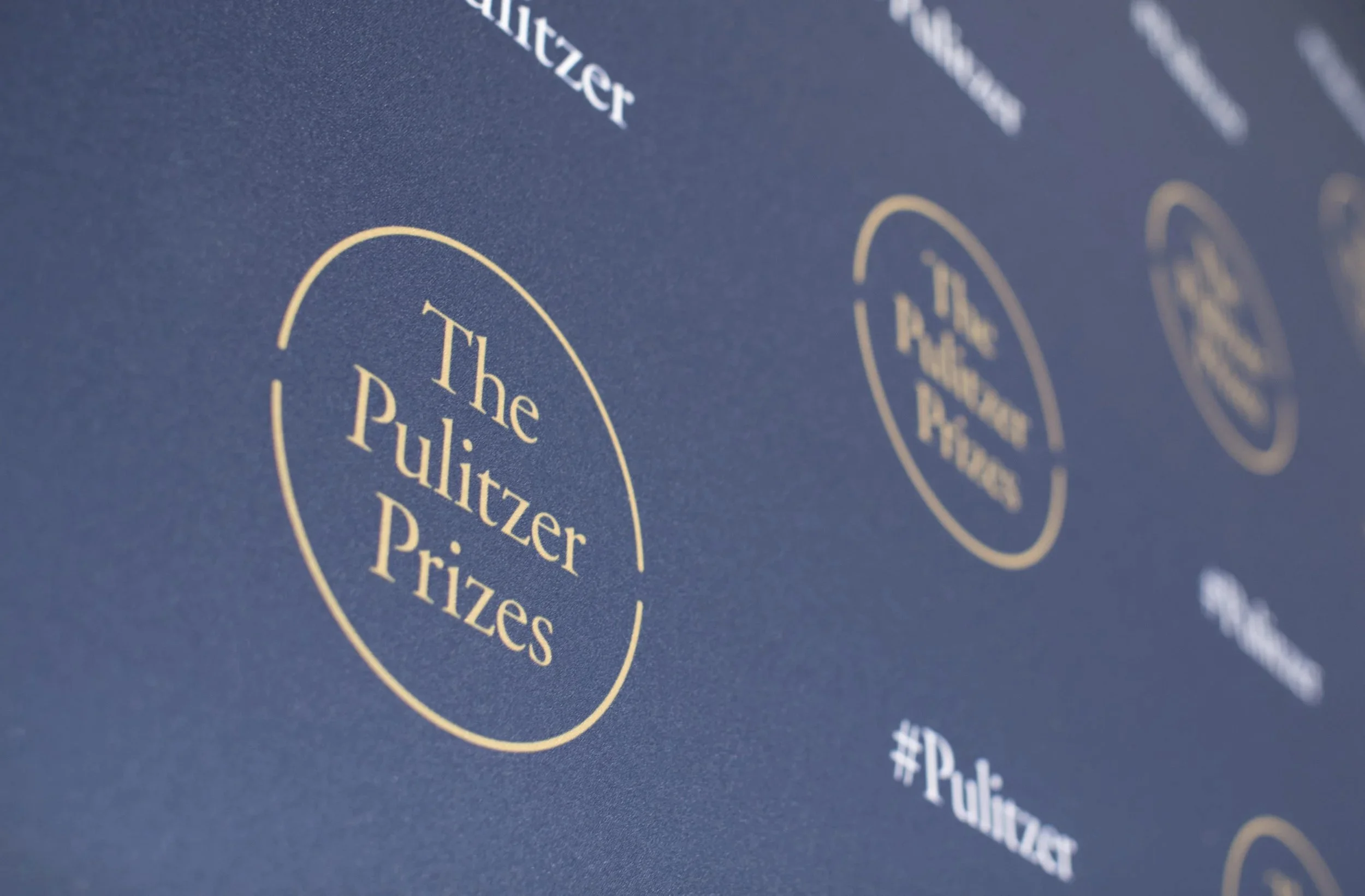 Close-up of a blue paper with gold and white text, featuring the words 'The Pulitzer Prizes' inside a gold circle.