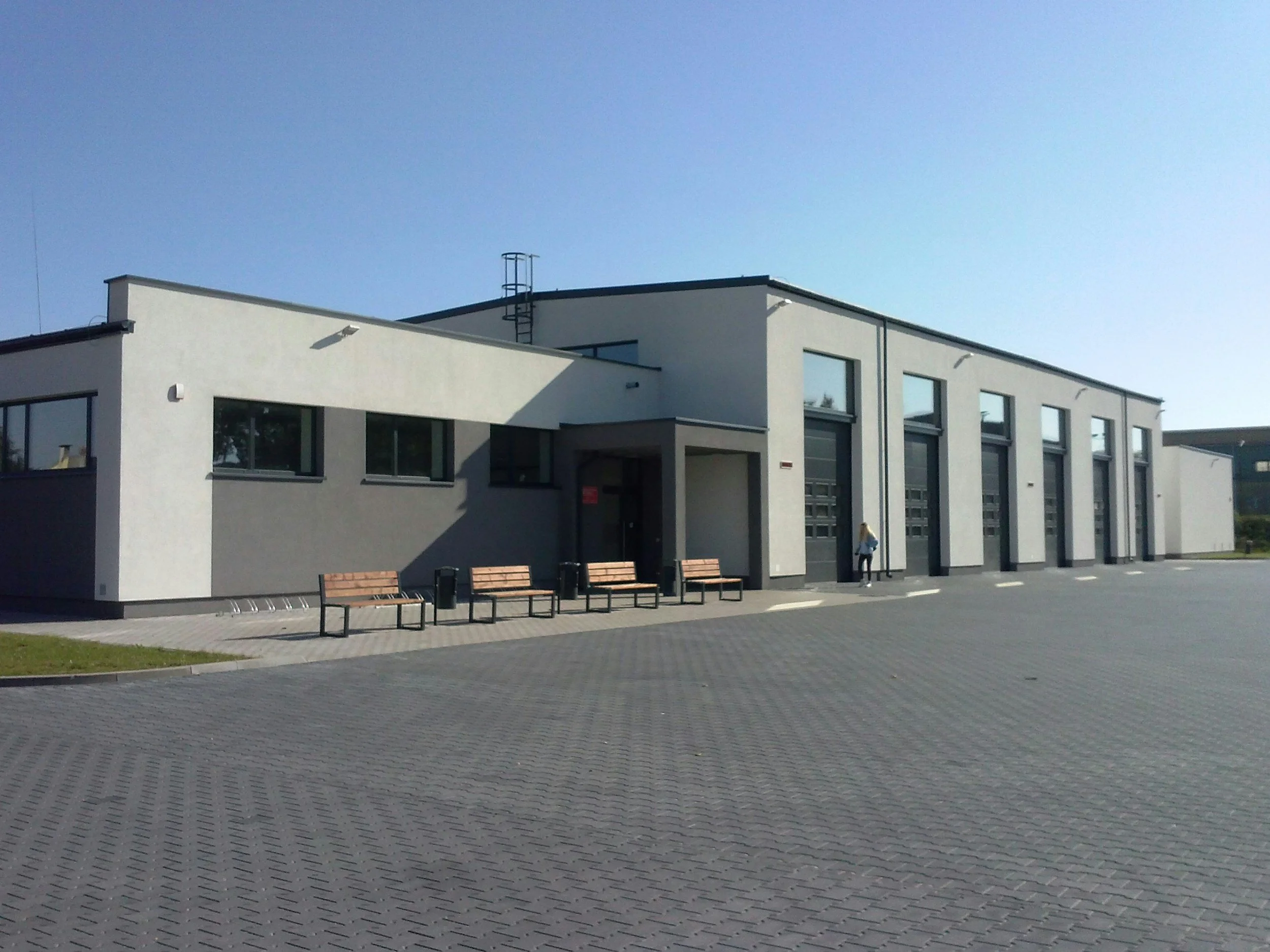 Modern white building with large windows, elevated on stilts, with a small person standing near the entrance, benches outside, under a clear blue sky, paved area in front.