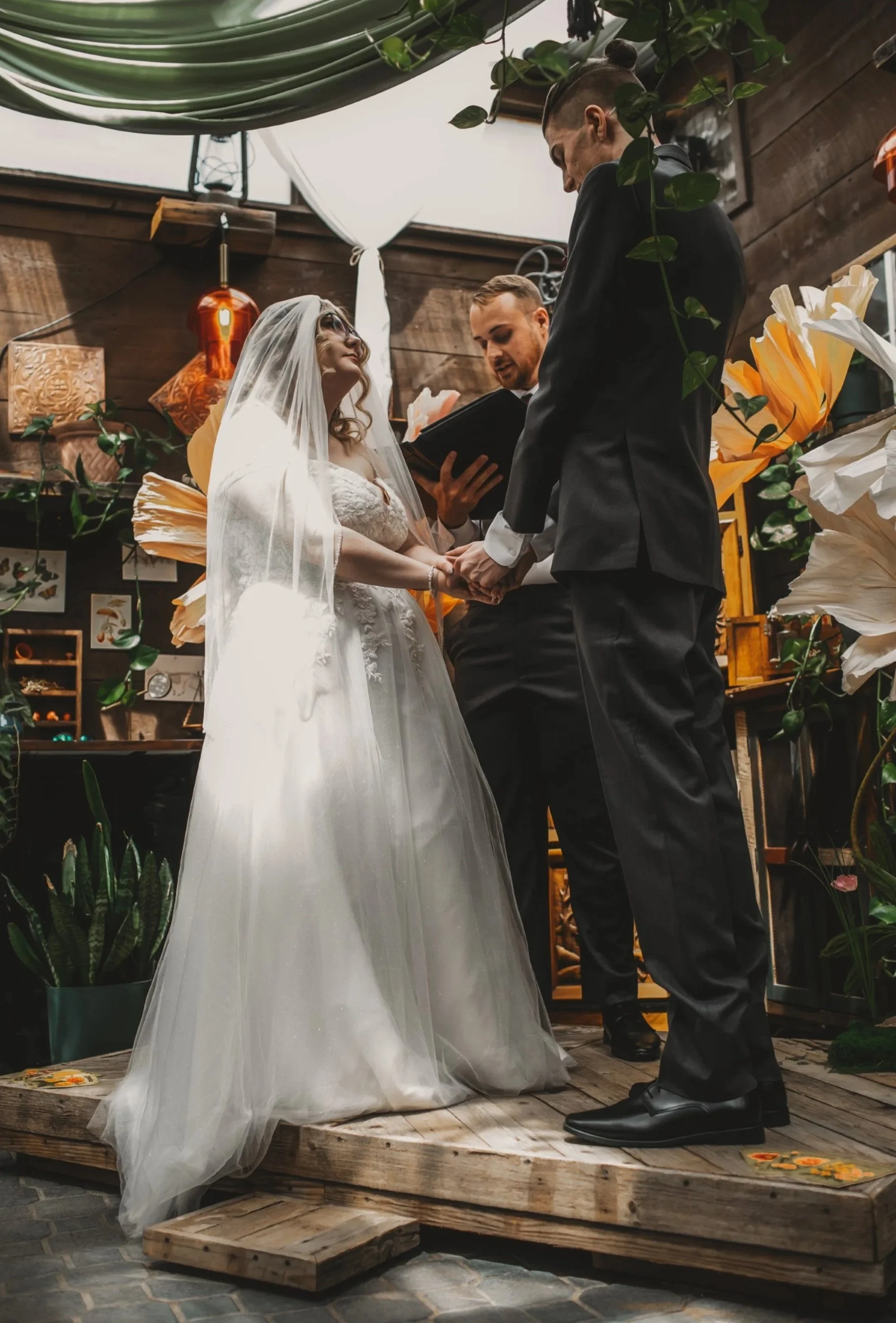 A bride and groom holding hands and exchanging vows during their wedding ceremony, with an officiant reading from a book, inside a rustic wooden venue decorated with plants and paper lanterns.