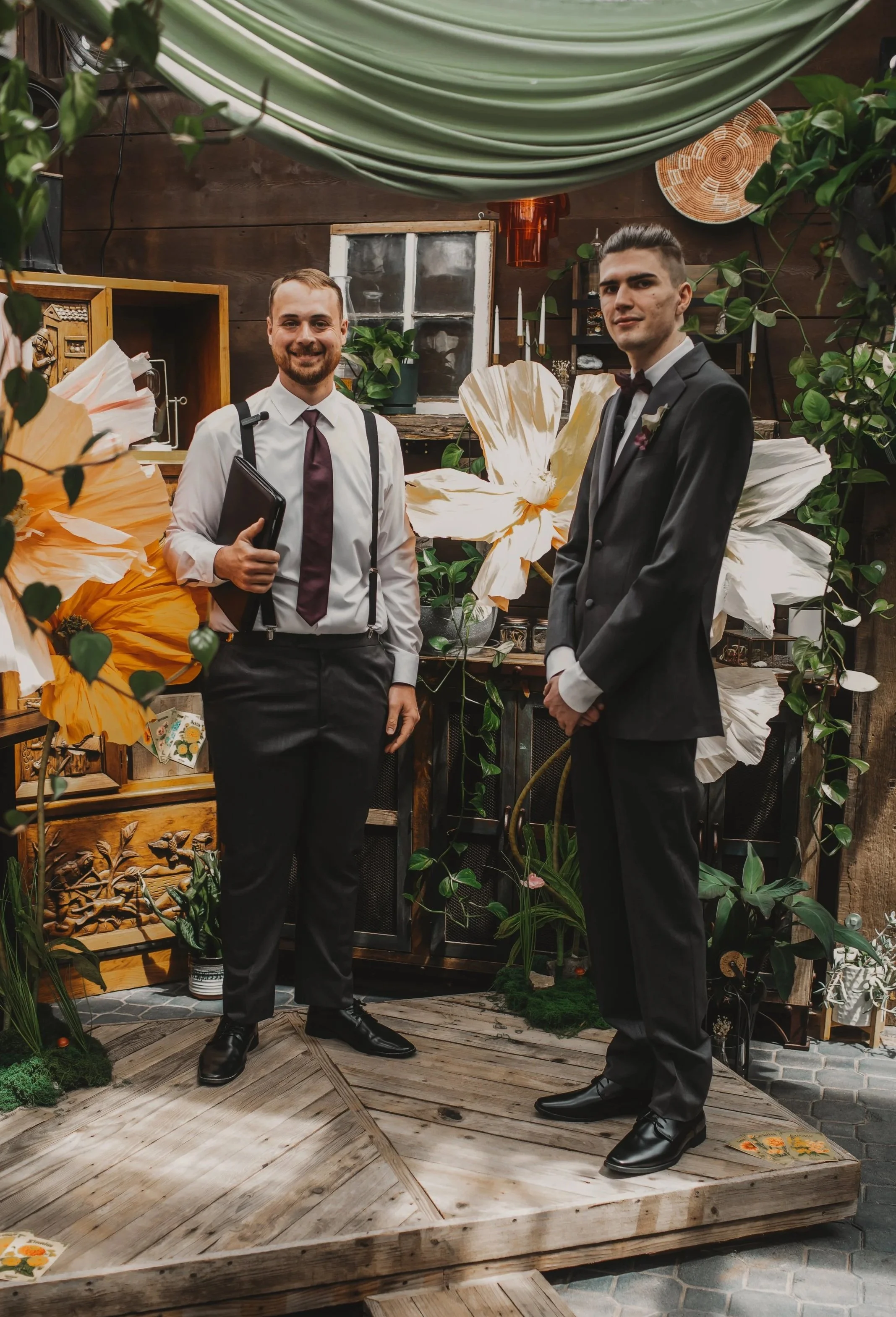 Two men in formal attire standing on a wooden platform amidst green plants and large decorative flowers, with a dark wooden wall and a small window in the background.