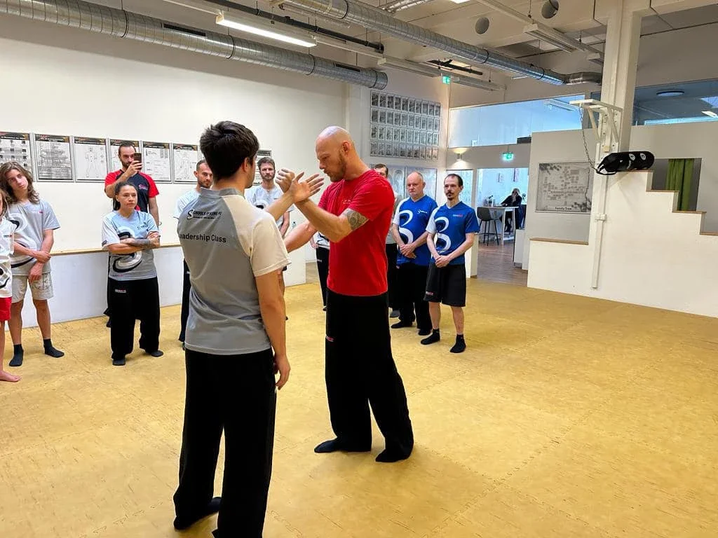 A martial arts or self-defense class with an instructor demonstrating a technique to a student, surrounded by other students observing indoors.