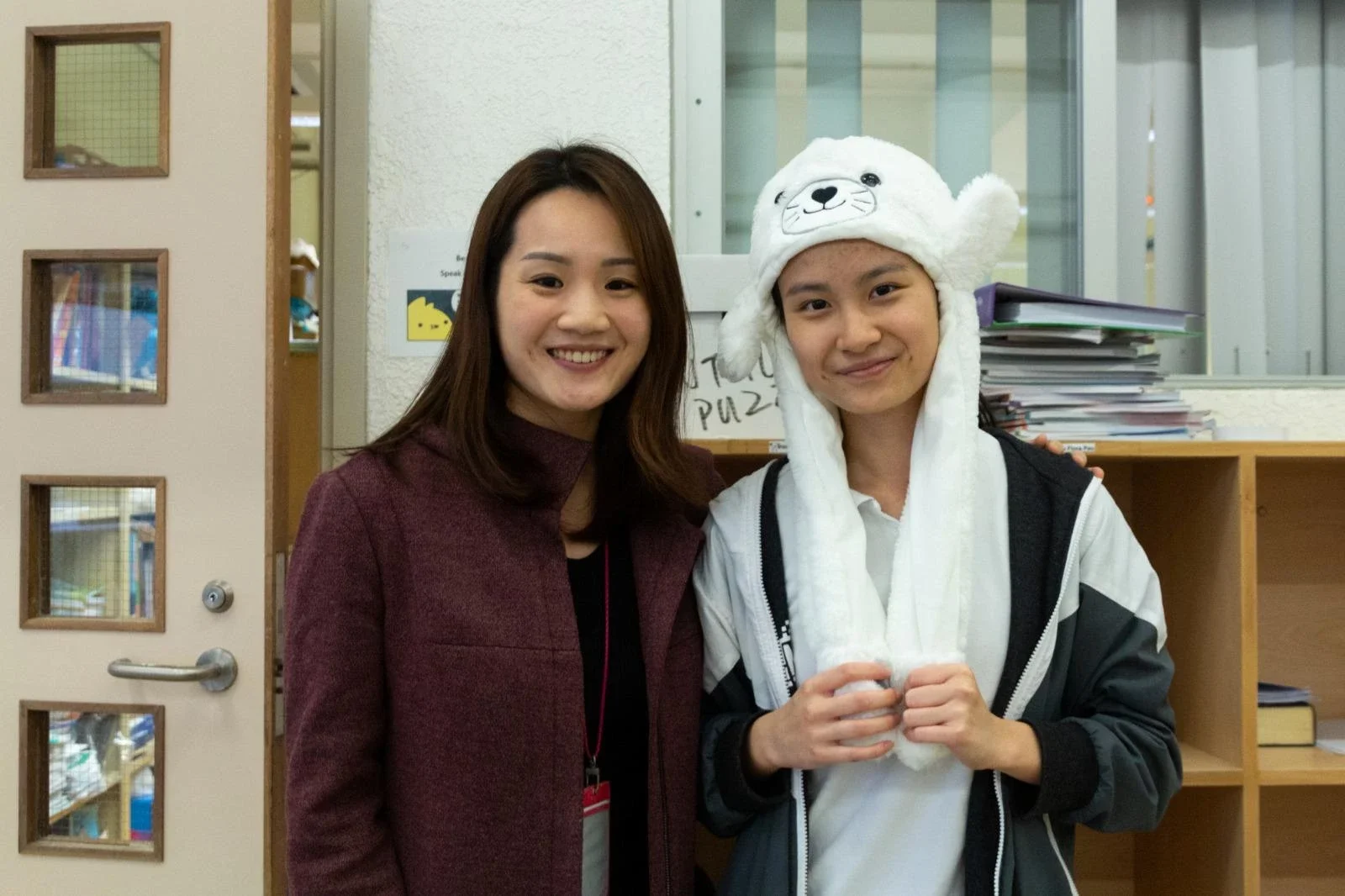Two women standing indoors, smiling. One woman is wearing a maroon jacket, and the other is wearing a white hat with a bear face, a white scarf, and a black jacket.