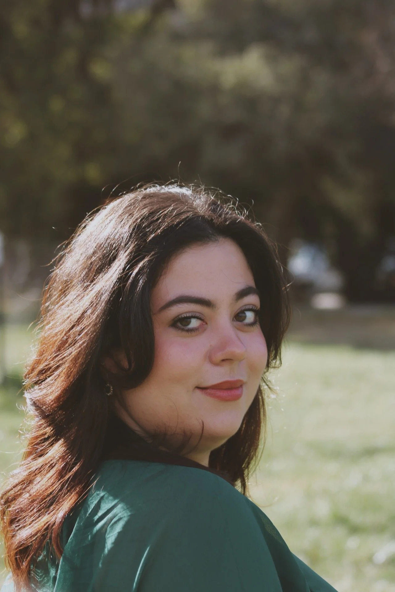 Isabella looking over her shoulder outdoors in a park with trees in the background.