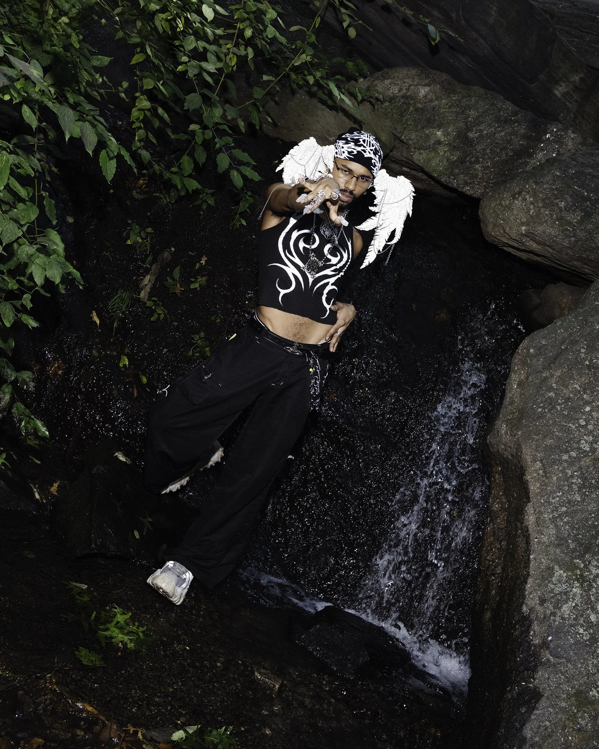 A person with dark hair, glasses, and an angel wing head piece standing in a rocky creek with water, surrounded by green plants and rocks, making a hand gesture towards the camera.