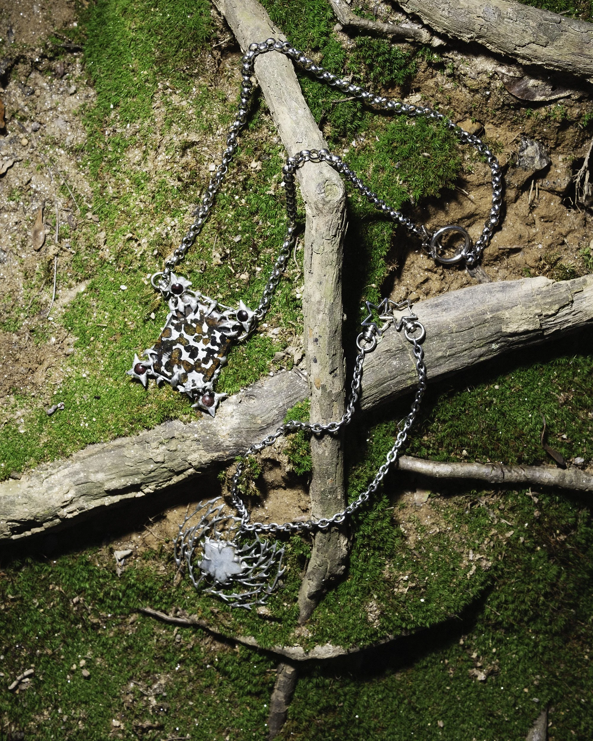 A silver necklace with star-shaped charms and a large floral pendant resting on a bed of moss, twigs, and soil outdoors.