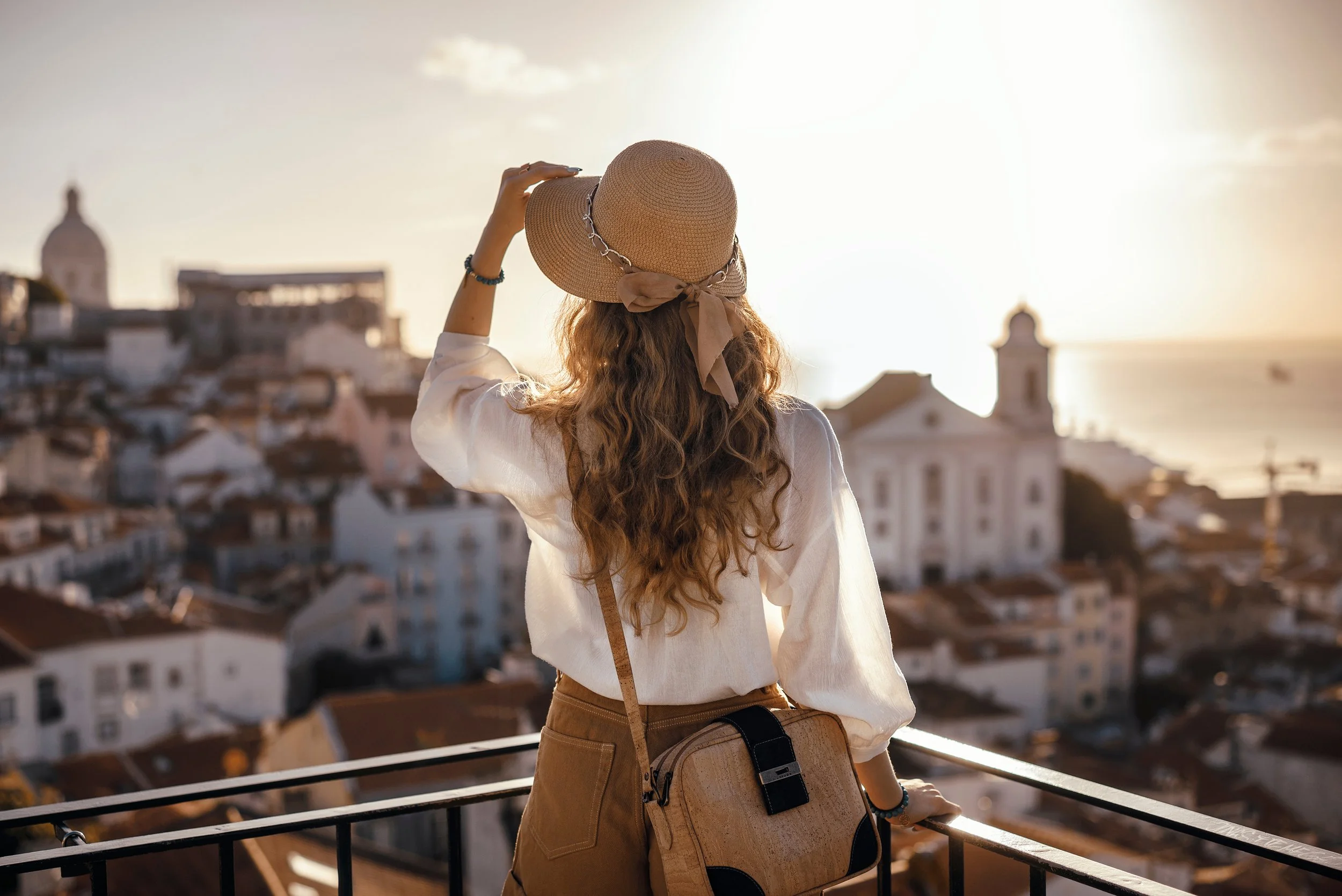 A woman with long, curly hair wearing a wide-brimmed straw hat and a white blouse looks at a cityscape at sunset, with buildings and the ocean in the background.