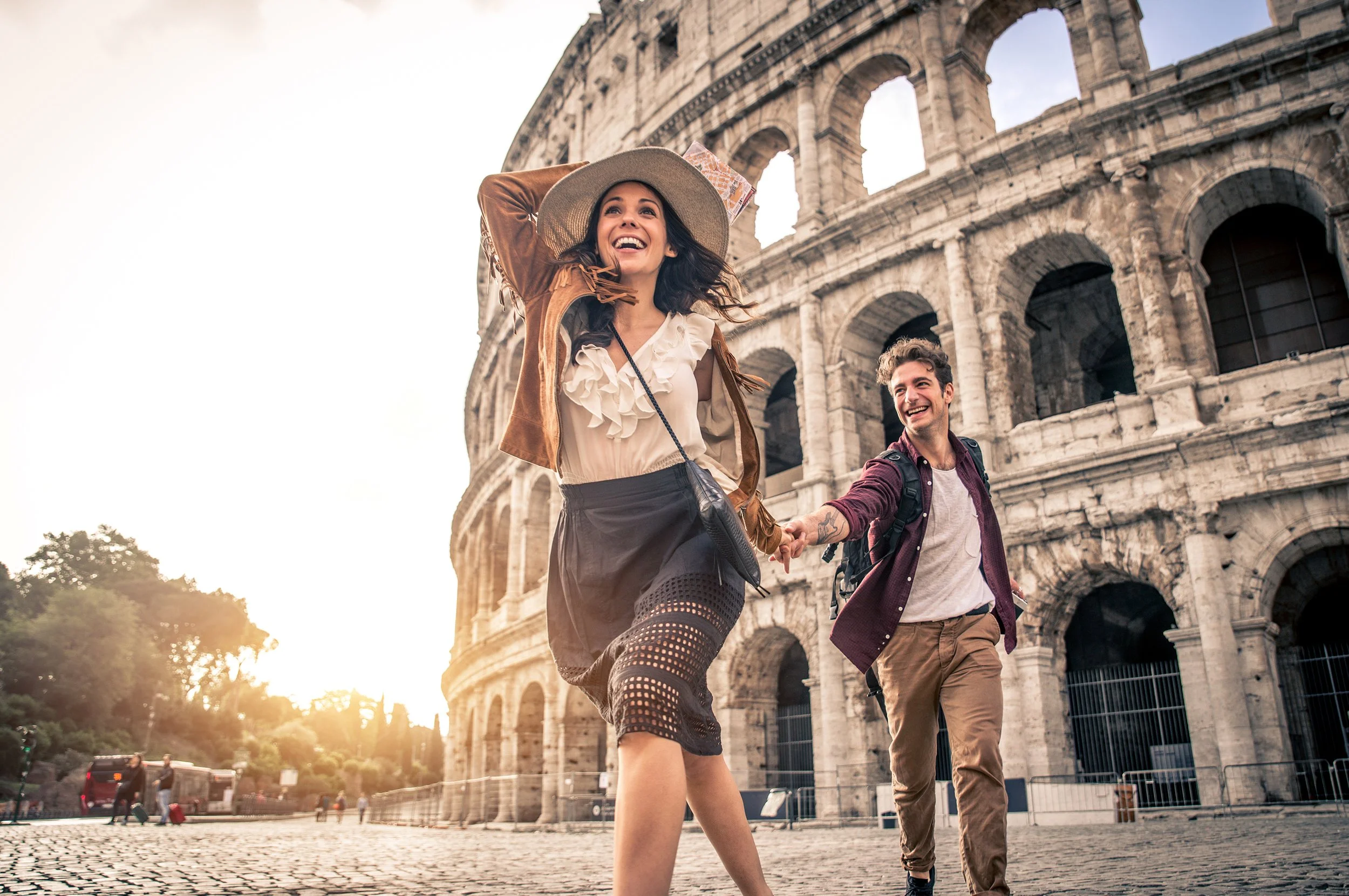 A woman and a man holding hands and smiling near the Roman Colosseum at sunset.