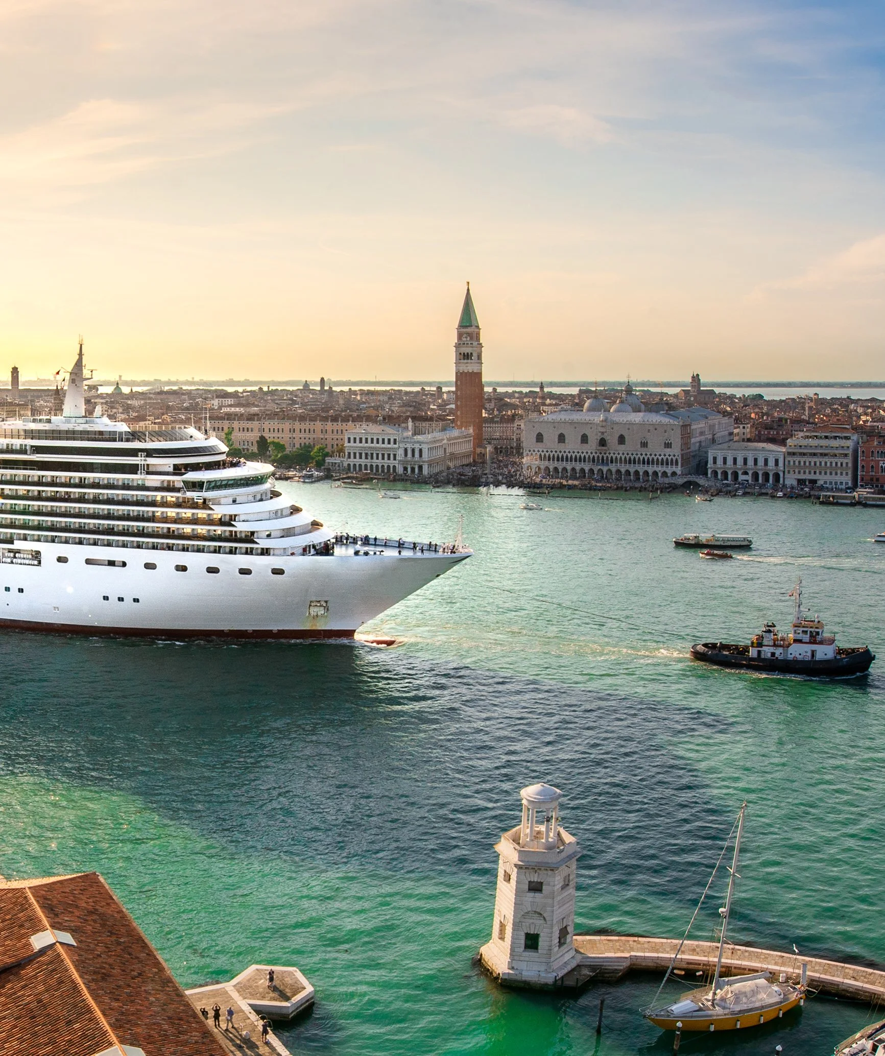 A view of Venice, Italy, showing a large cruise ship docked near a historic tower and buildings along the water. Small boats and people are visible around the waterfront.