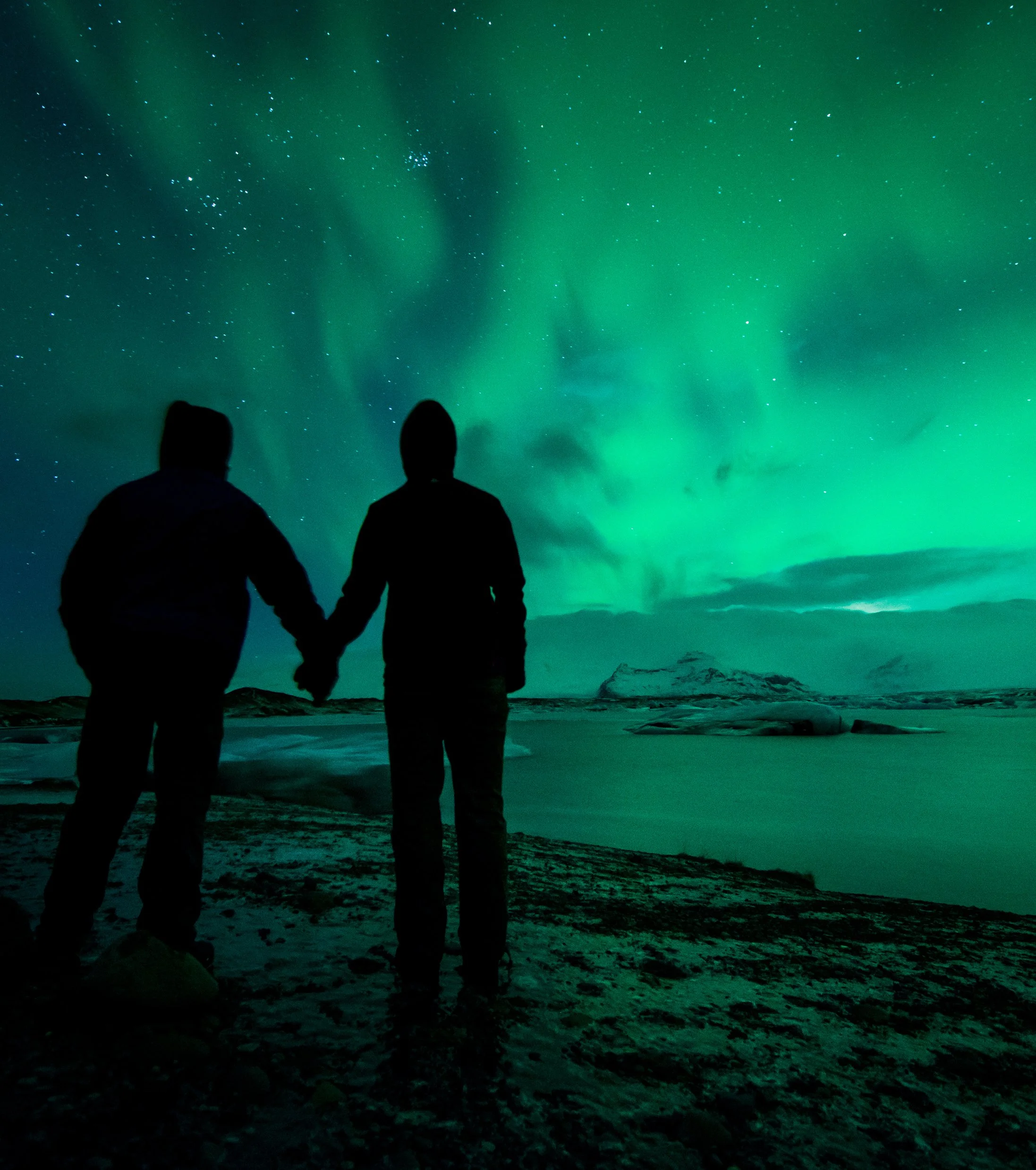 Two silhouetted people holding hands on a rocky terrain, gazing at the night sky illuminated by green northern lights and starry sky.
