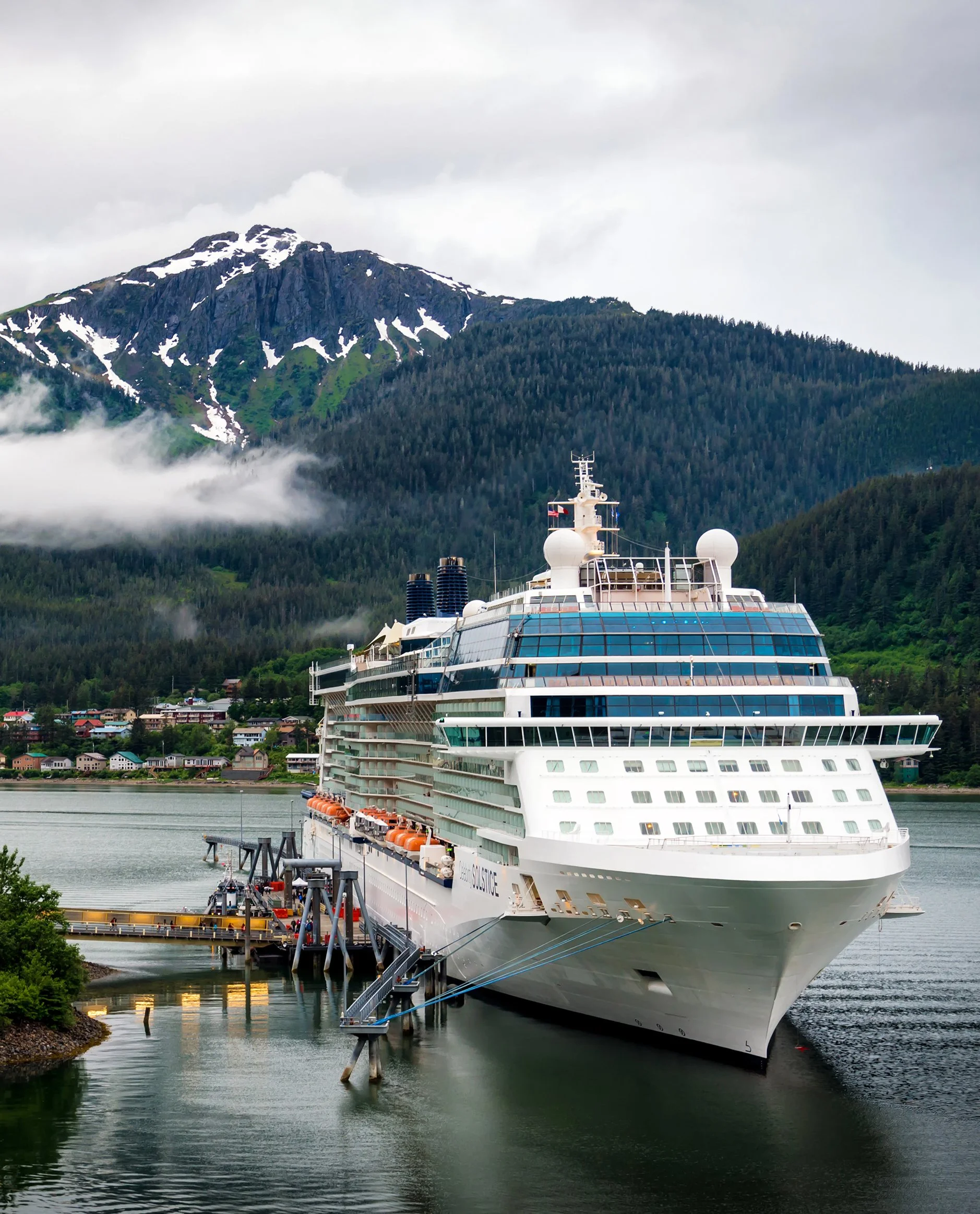 A large white cruise ship docked at a port with a mountainous landscape in the background, partially covered with snow and greenery.