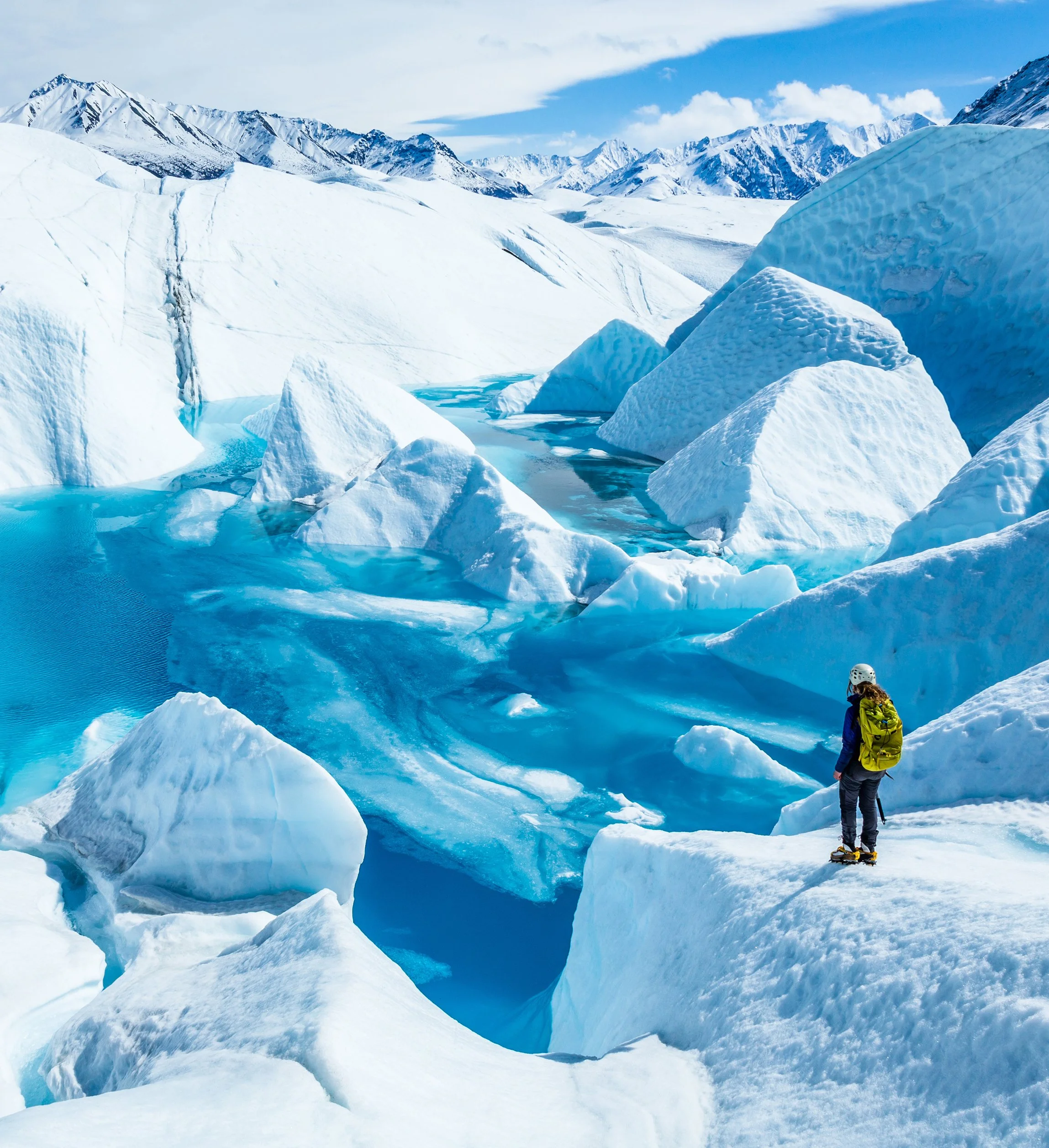 A person standing on a yellow skateboard with a green backpack in Alaska, navigating over ice and snow in a glacial landscape, with snow-covered mountains in the background and a partly cloudy sky.
