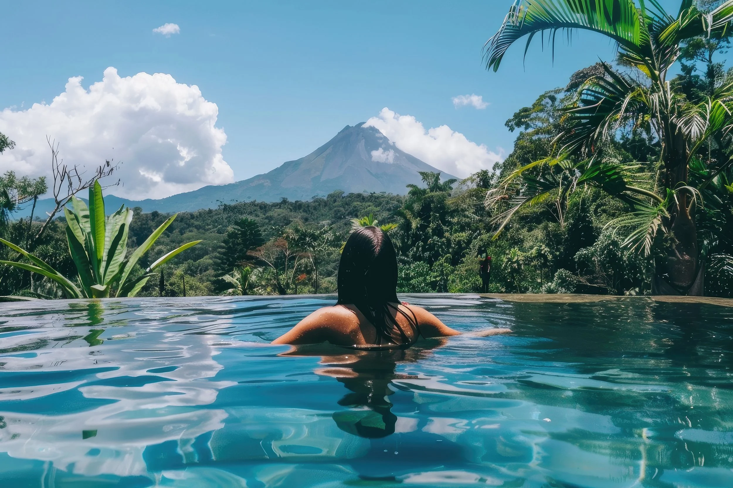 A person with wet hair relaxing in an infinity pool with a mountain and lush tropical trees in the background.
