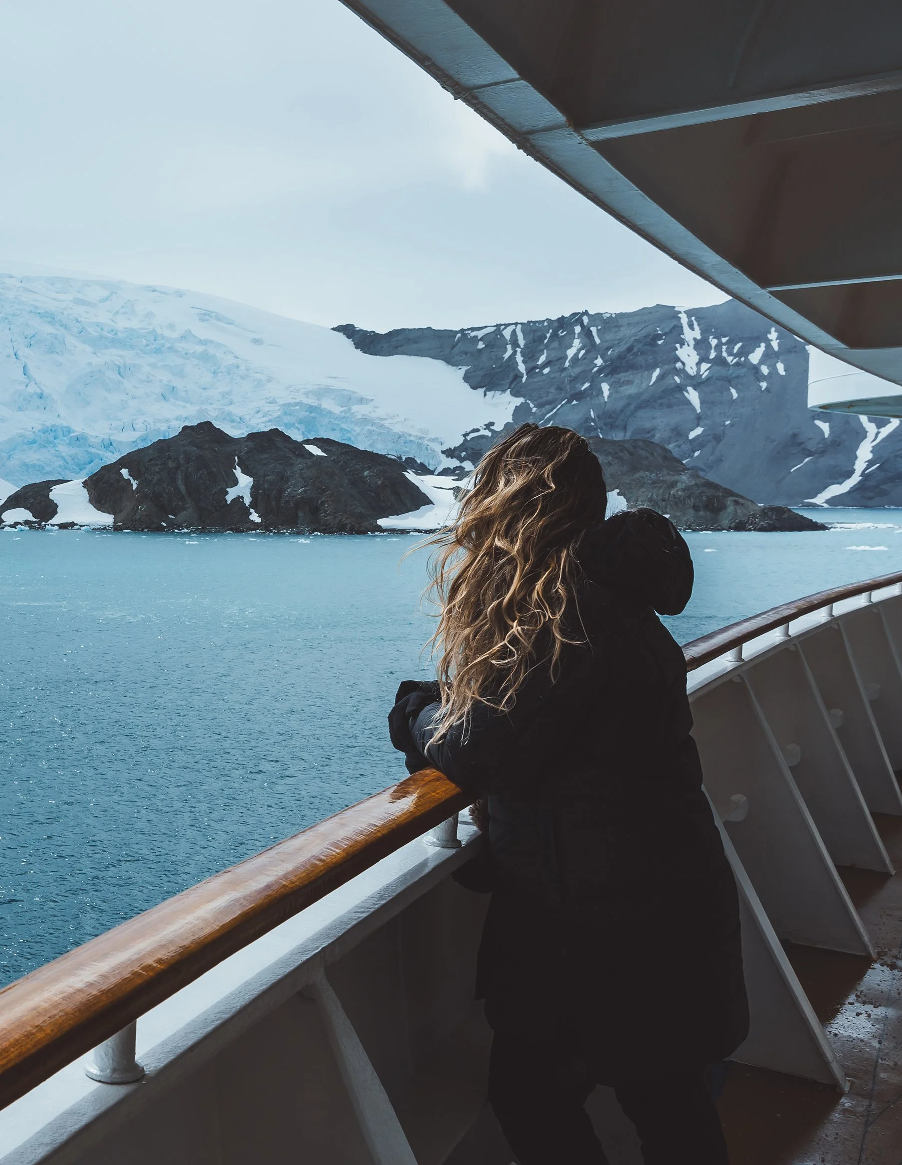 A person with long, wavy hair standing on a ship balcony, looking out at icy mountains and a glacier in the distance.