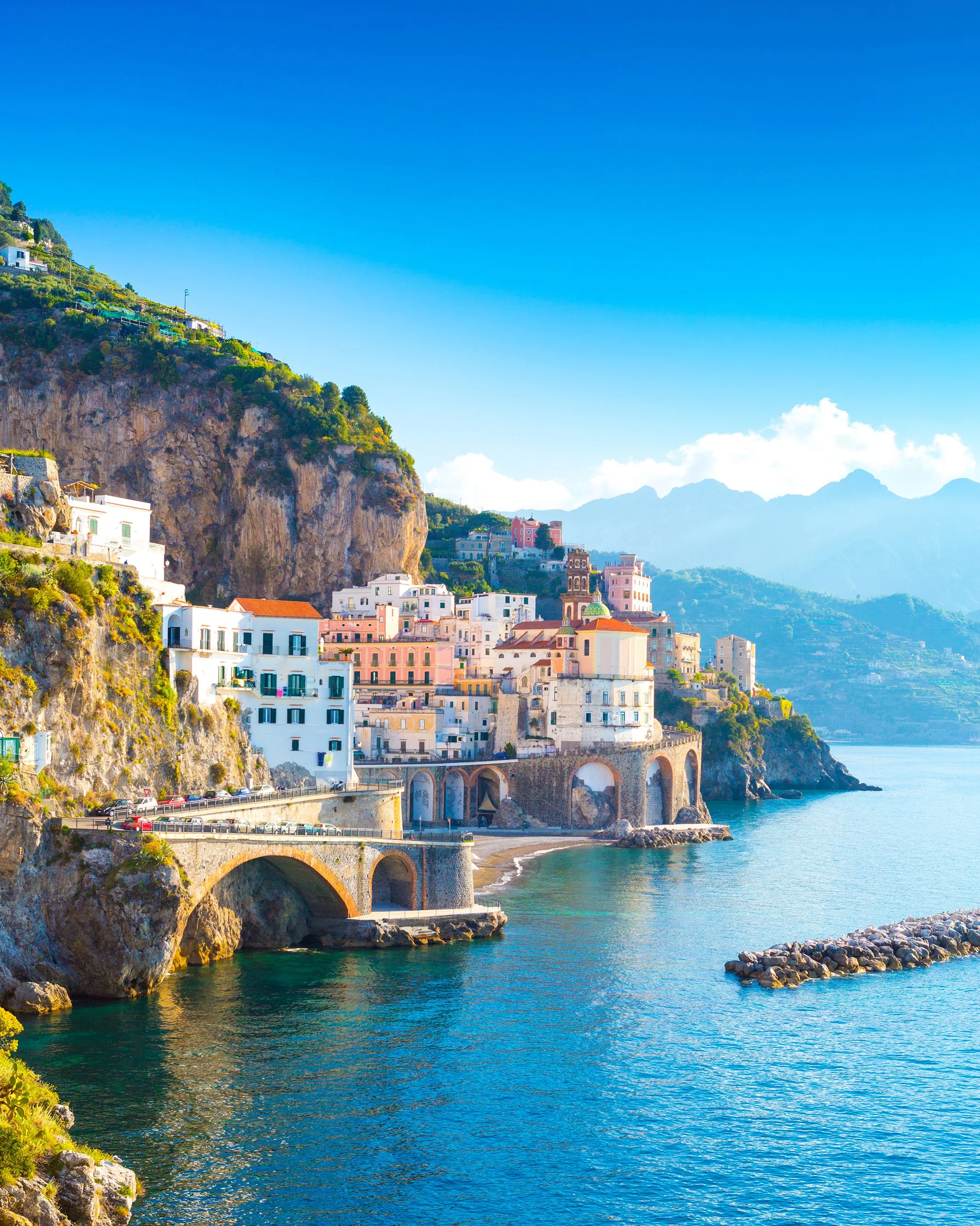Colorful buildings on a coastal hillside with a bridge over turquoise water and distant mountains in clear sky.