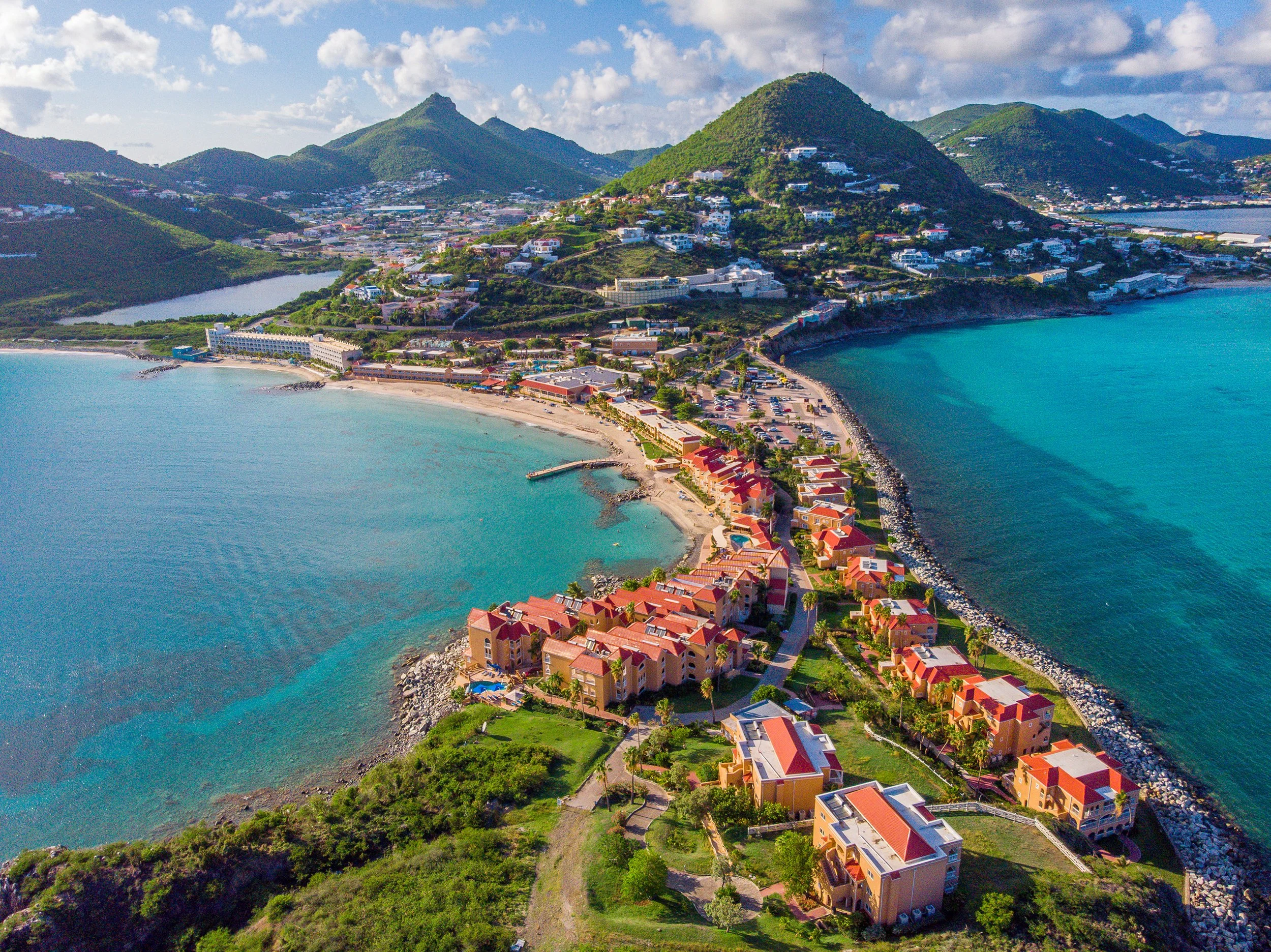 Aerial view of a coastal town with colorful buildings, mountains covered in green vegetation, and blue ocean water.