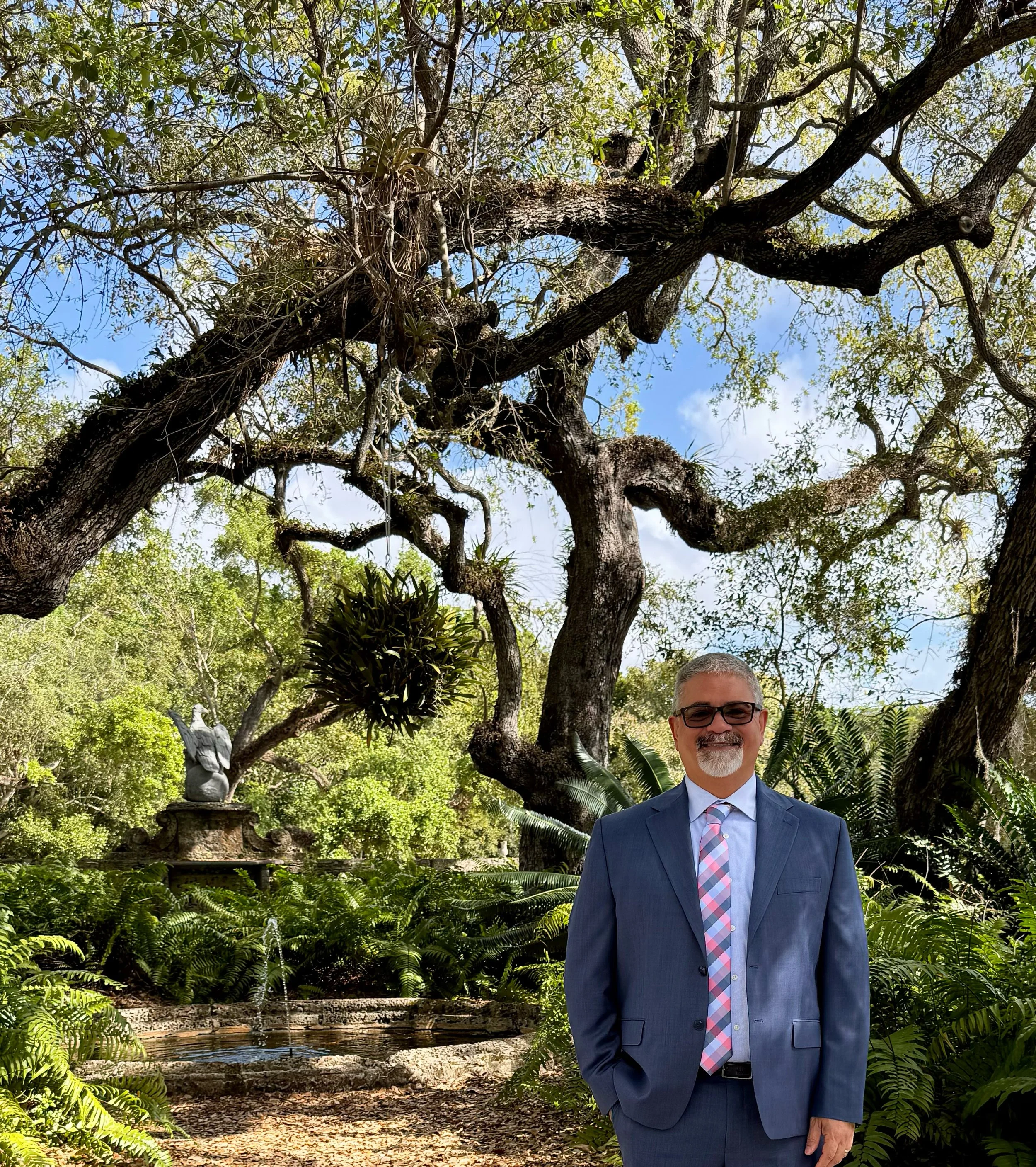A man in a blue suit with a patterned tie stands outdoors in front of lush green trees and plants. The background features a small pond with a fountain, a statue, and a fountain stone. The scene is bright and sunny, with clear blue sky visible through the tree leaves.