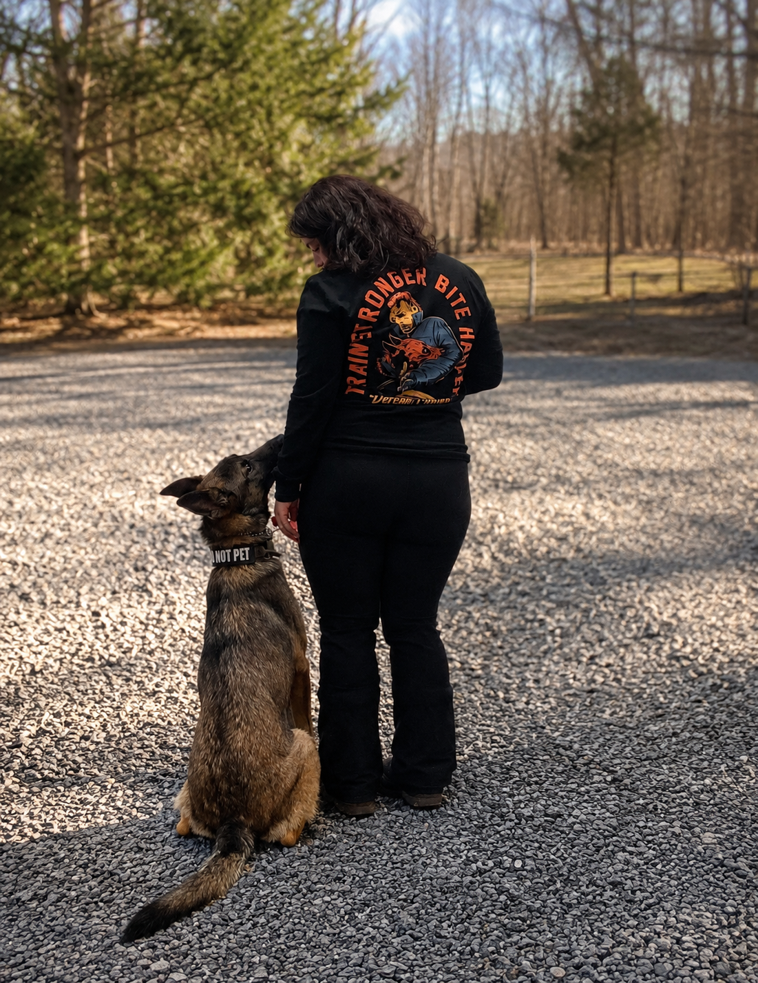 A woman standing outdoors on a gravel surface with a German Shepherd dog sitting beside her. The woman is wearing a black jacket with an 'Overlord Biker' patch on the back, and the dog has a collar that reads 'I AM NOT PET.' In the background, there are trees with some greenery and some no leaves, and a sunny sky.