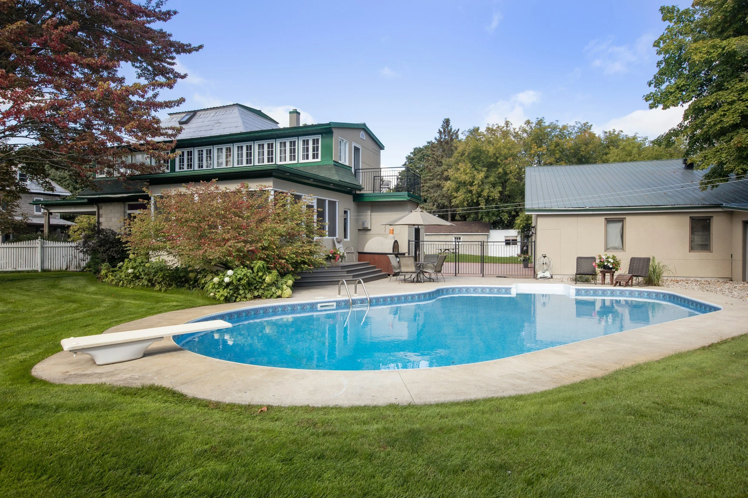 A backyard with a swimming pool, surrounded by green grass, trees, and outdoor furniture. There is a house with a patio and a grill in the background under a partly cloudy sky.