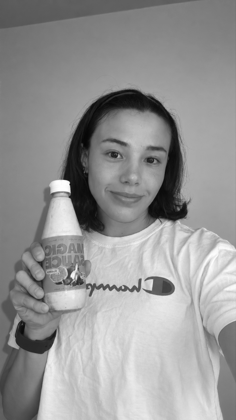 Mujer joven con cabello castaño oscuro y camiseta blanca sosteniendo una botella de jugo de cítricos y ajo, sonriendo, en un fondo de pared blanca.