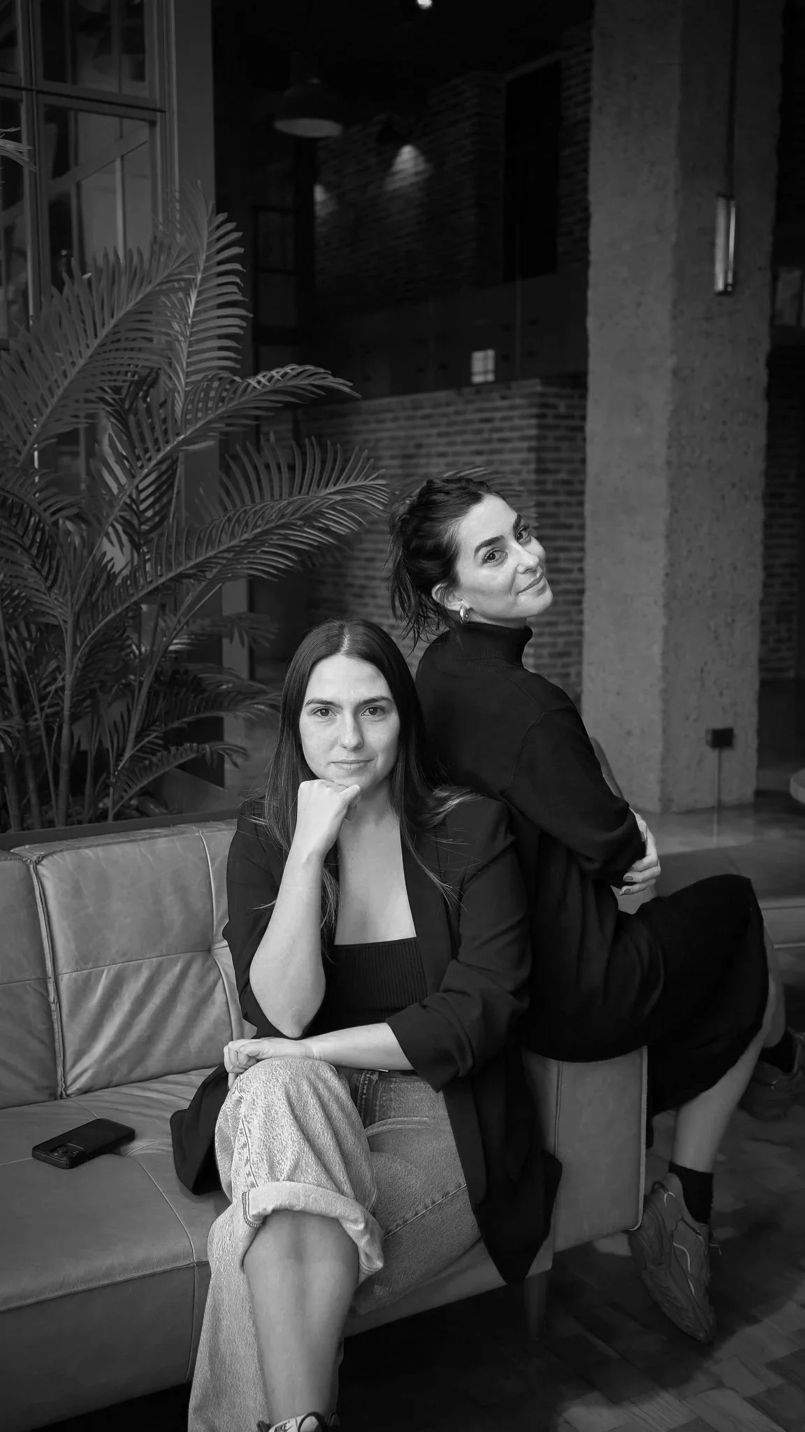 Dos mujeres sentadas en un sofá en un ambiente interior, una de ellas con el cabello largo y la otra con el cabello recogido, ambas con expresión tranquila.