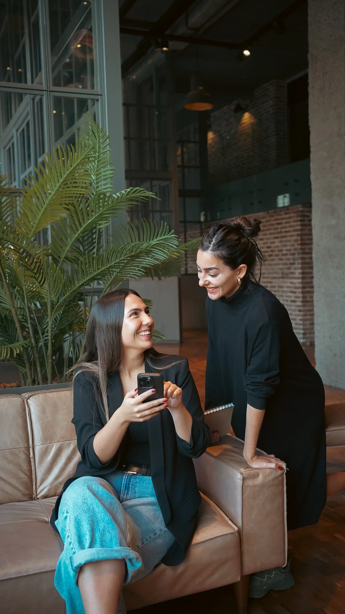 Dos mujeres en un ambiente interior acogedor, una sentada usando un teléfono y la otra de pie, interactuando; fondo con plantas y paredes de ladrillo.